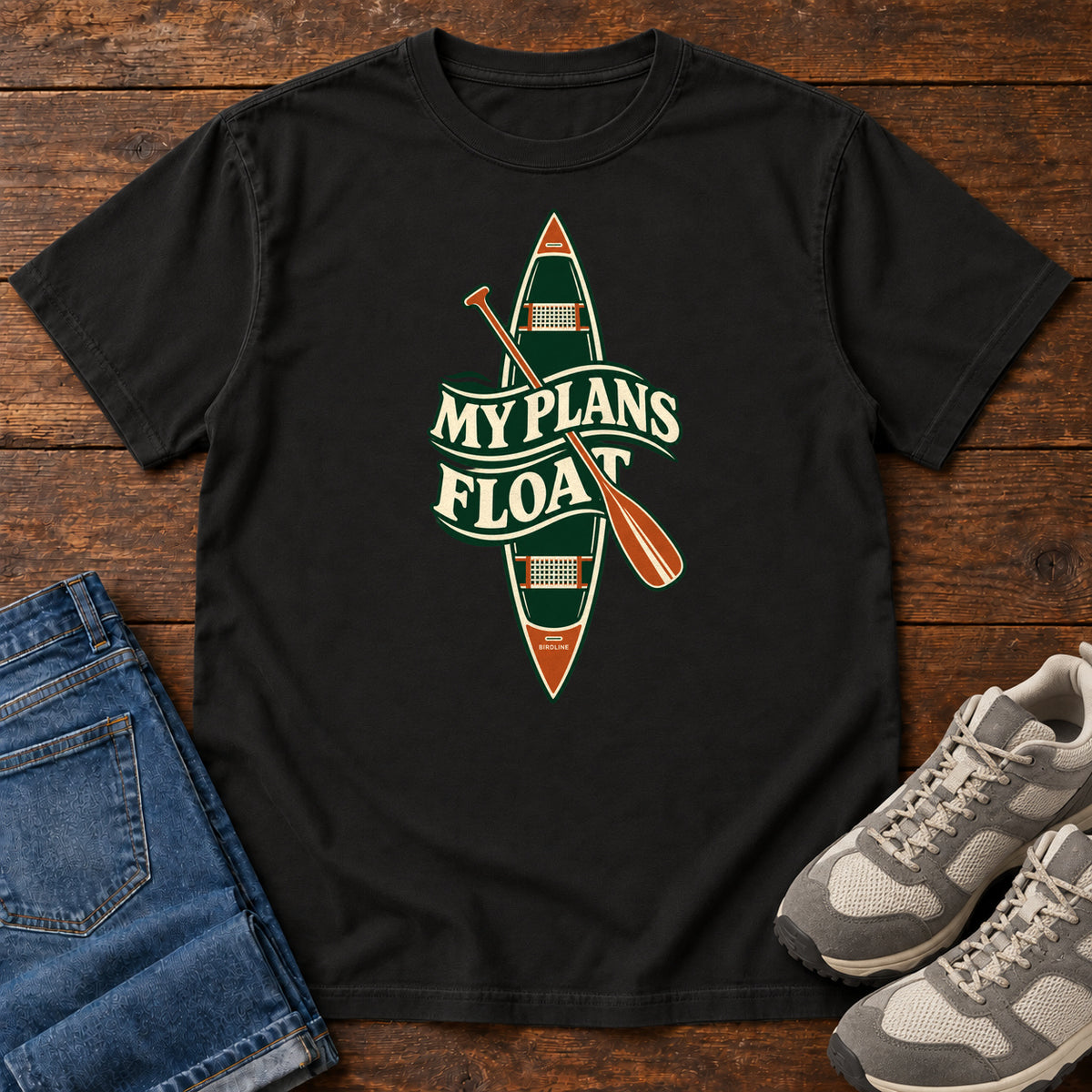 My Plans Float T-Shirt in Black, front laid flat on wood, featuring a vertical green canoe and orange paddle behind cream slogan lettering.