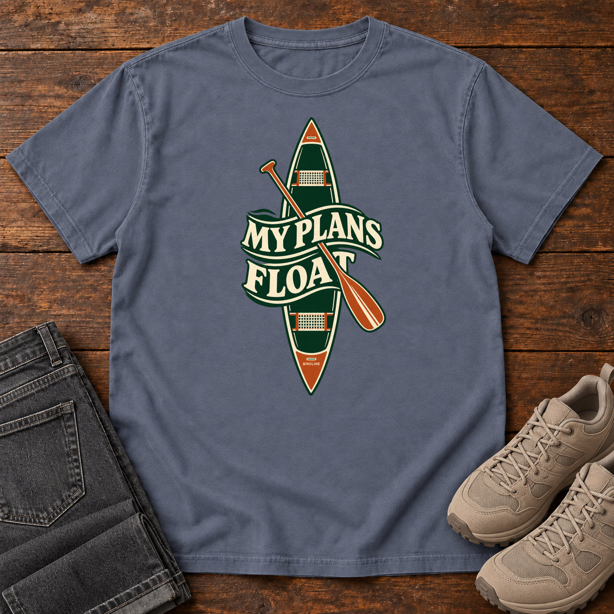 My Plans Float T-Shirt in Denim, front laid flat on wood, featuring a vertical green canoe and orange paddle behind cream slogan lettering.