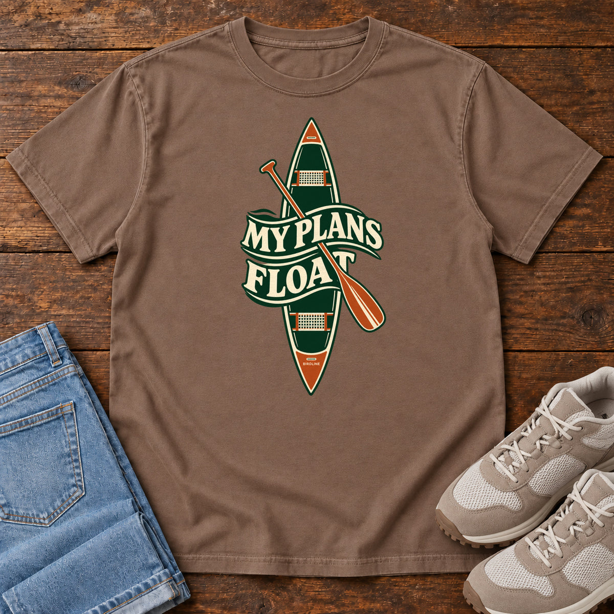 My Plans Float T-Shirt in Espresso, front laid flat on wood, featuring a vertical green canoe and orange paddle behind cream slogan lettering.