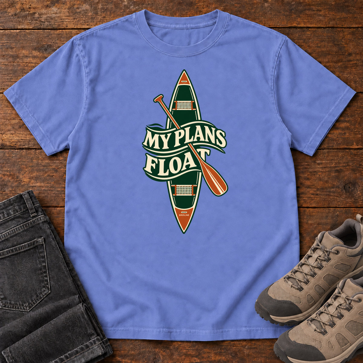 My Plans Float T-Shirt in Flo Blue, front laid flat on wood, featuring a vertical green canoe and orange paddle behind cream slogan lettering.