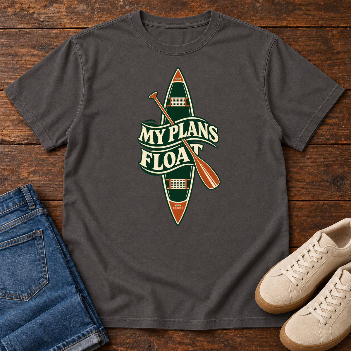 My Plans Float T-Shirt in Graphite, front laid flat on wood, featuring a vertical green canoe and orange paddle behind cream slogan lettering.