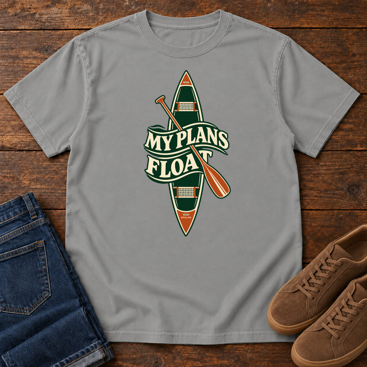 My Plans Float T-Shirt in Grey, front laid flat on wood, featuring a vertical green canoe and orange paddle behind cream slogan lettering.