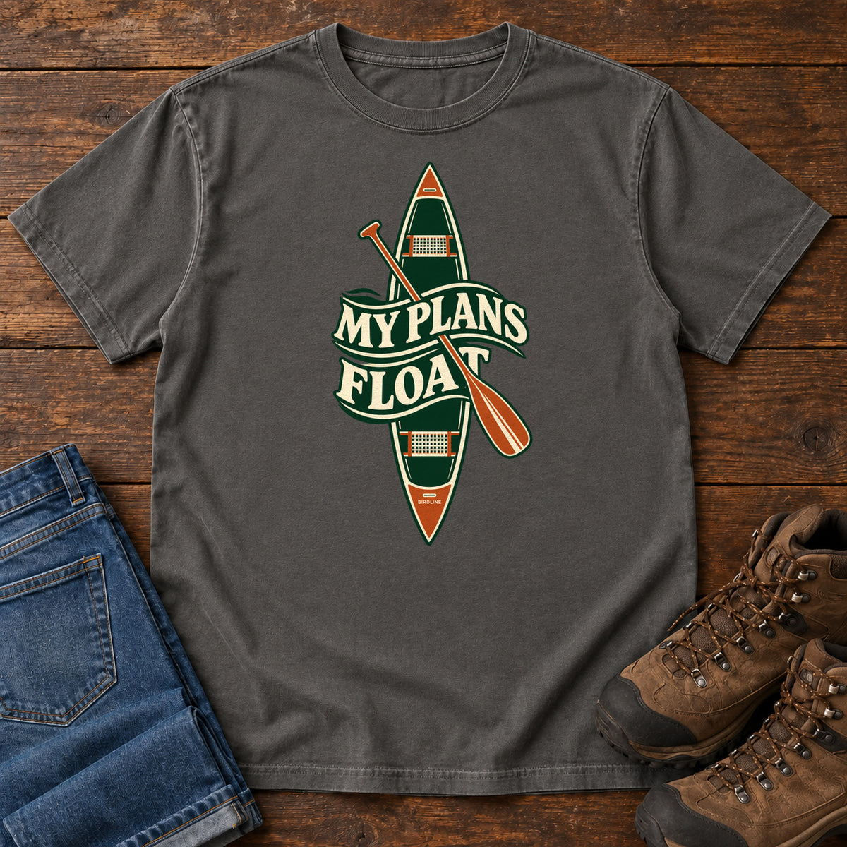 My Plans Float T-Shirt in Pepper, front laid flat on wood, featuring a vertical green canoe and orange paddle behind cream slogan lettering.
