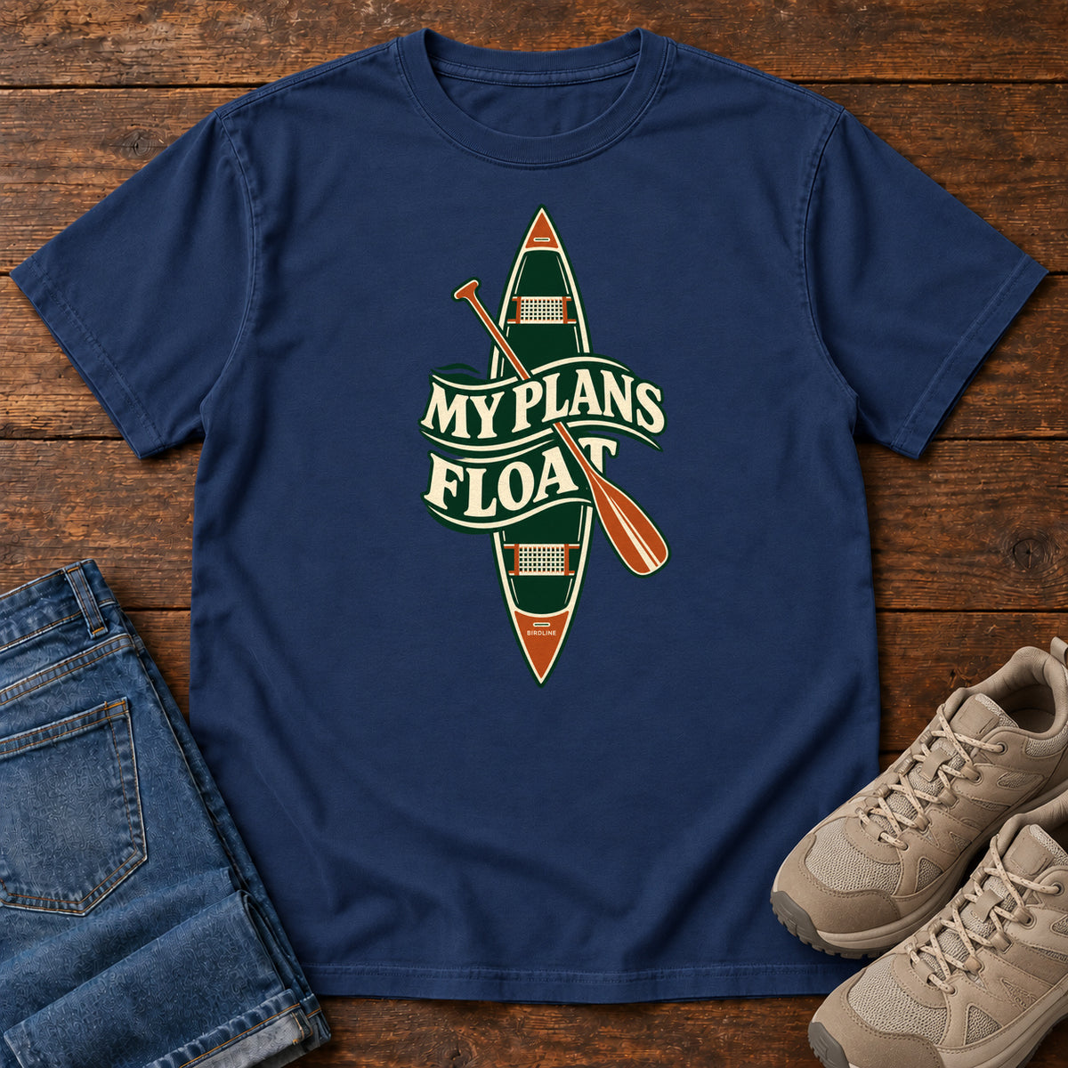 My Plans Float T-Shirt in True Navy, front laid flat on wood, featuring a vertical green canoe and orange paddle behind cream slogan lettering.