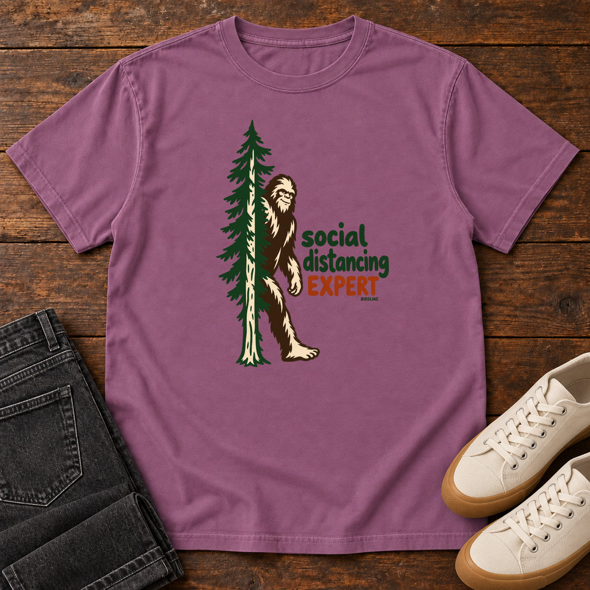 Berry Social Distancing Expert T-Shirt with Bigfoot beside an evergreen and Social Distancing Expert lettering; front flat lay on wood.