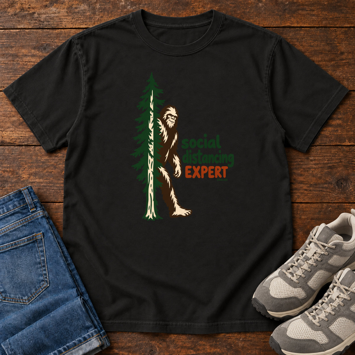Black Social Distancing Expert T-Shirt with Bigfoot beside an evergreen and Social Distancing Expert lettering; front flat lay on wood.