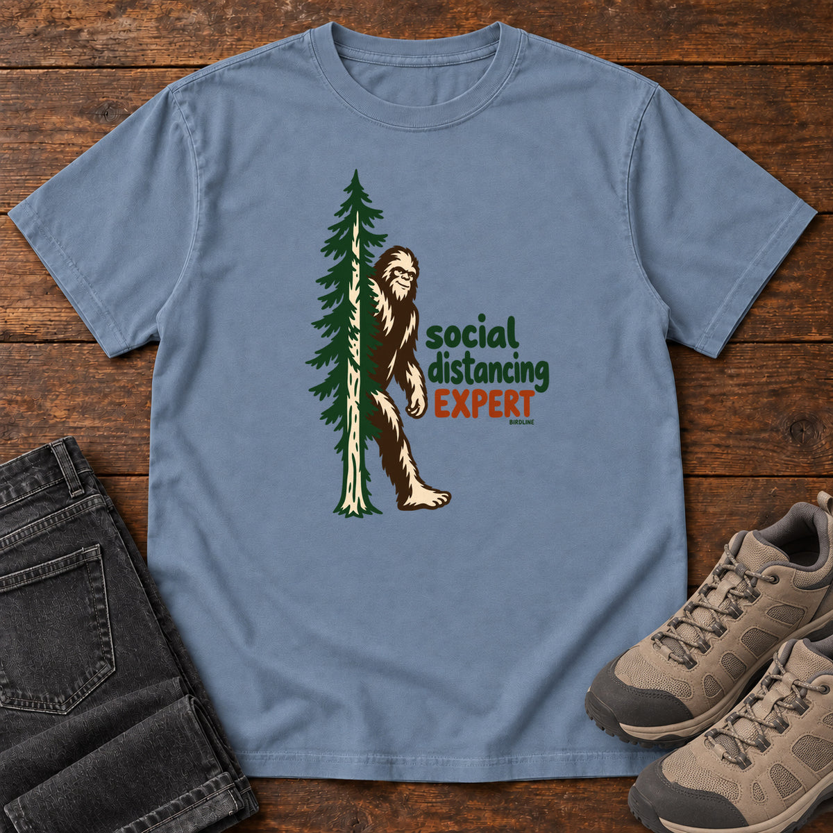 Blue Jean Social Distancing Expert T-Shirt with Bigfoot beside an evergreen and Social Distancing Expert lettering; front flat lay on wood.