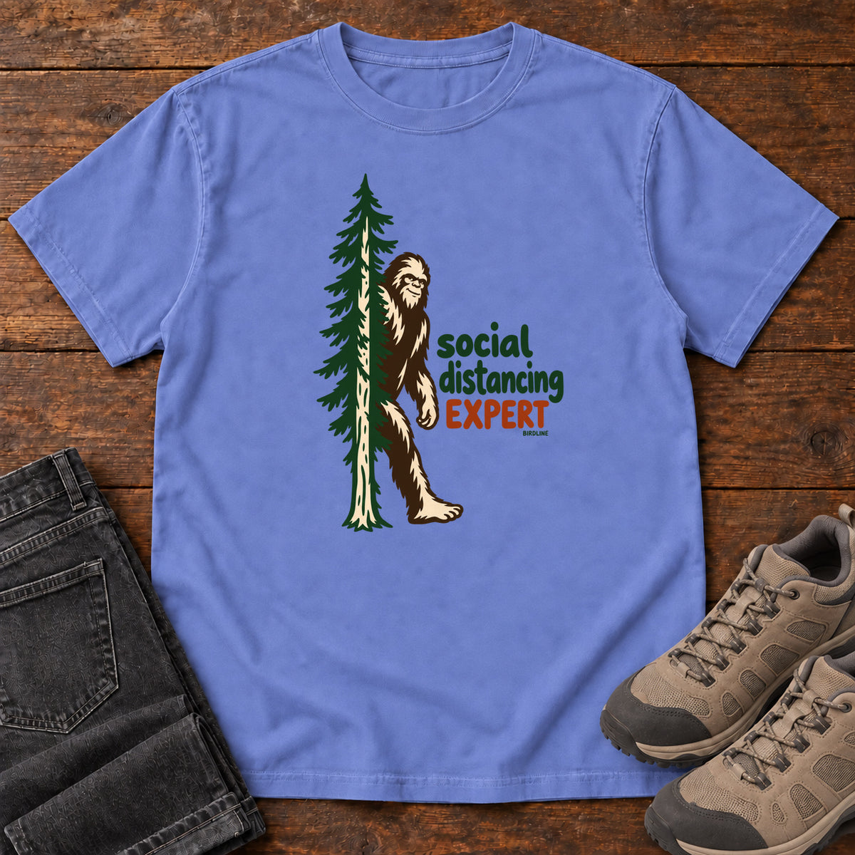 Flo Blue Social Distancing Expert T-Shirt with Bigfoot beside an evergreen and Social Distancing Expert lettering; front flat lay on wood.