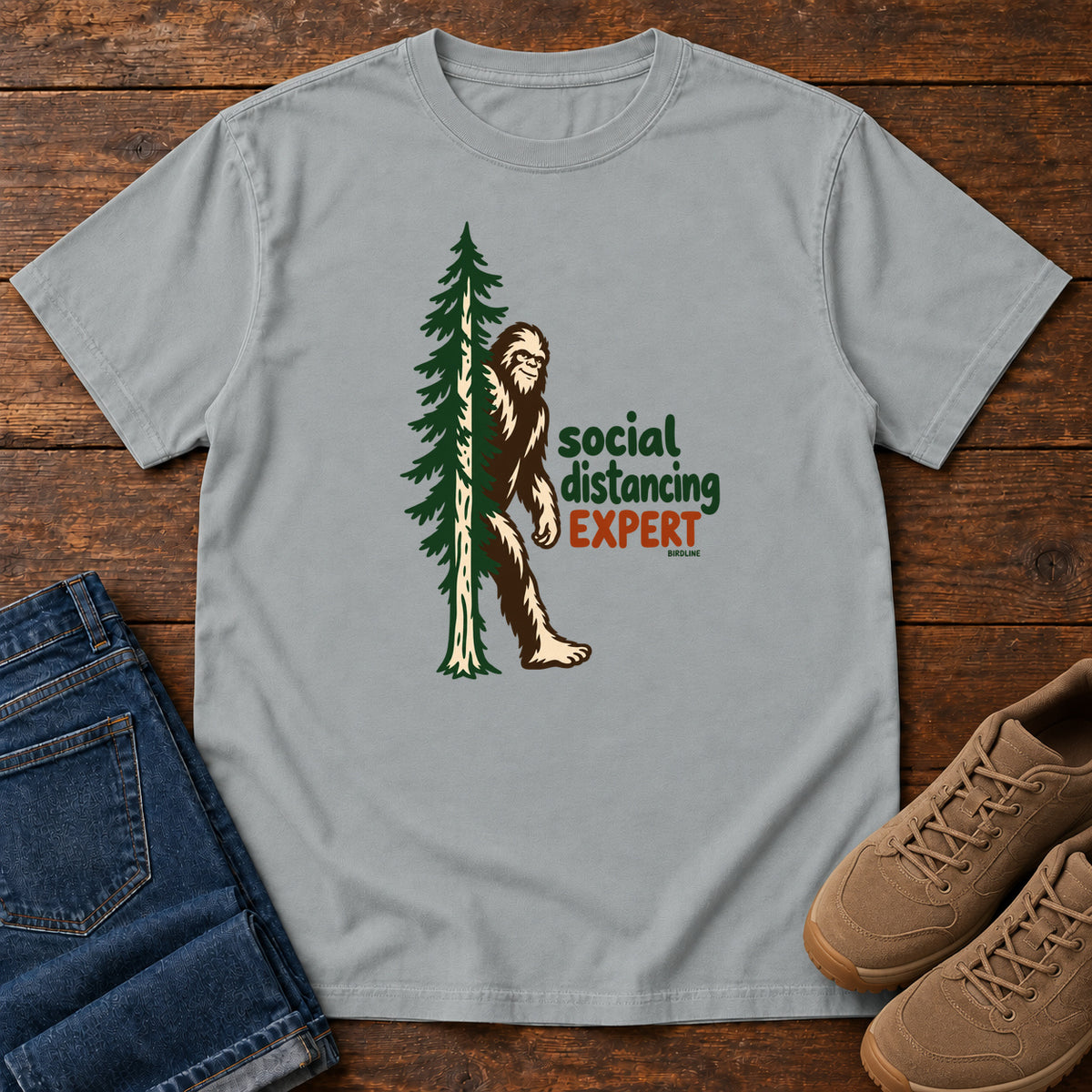Granite Social Distancing Expert T-Shirt with Bigfoot beside an evergreen and Social Distancing Expert lettering; front flat lay on wood.