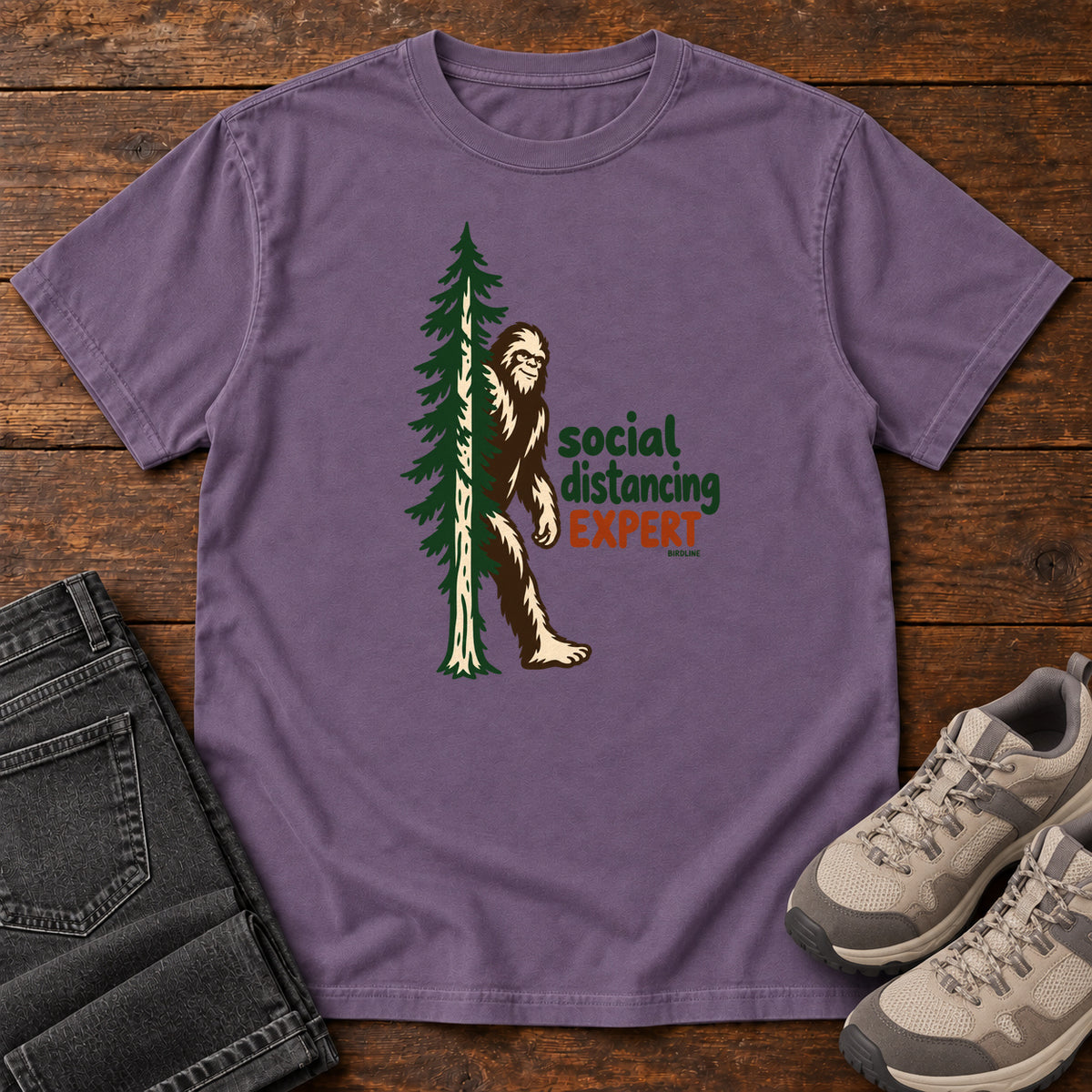 Grape Social Distancing Expert T-Shirt with Bigfoot beside an evergreen and Social Distancing Expert lettering; front flat lay on wood.