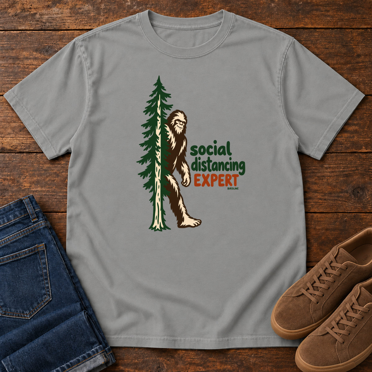 Grey Social Distancing Expert T-Shirt with Bigfoot beside an evergreen and Social Distancing Expert lettering; front flat lay on wood.