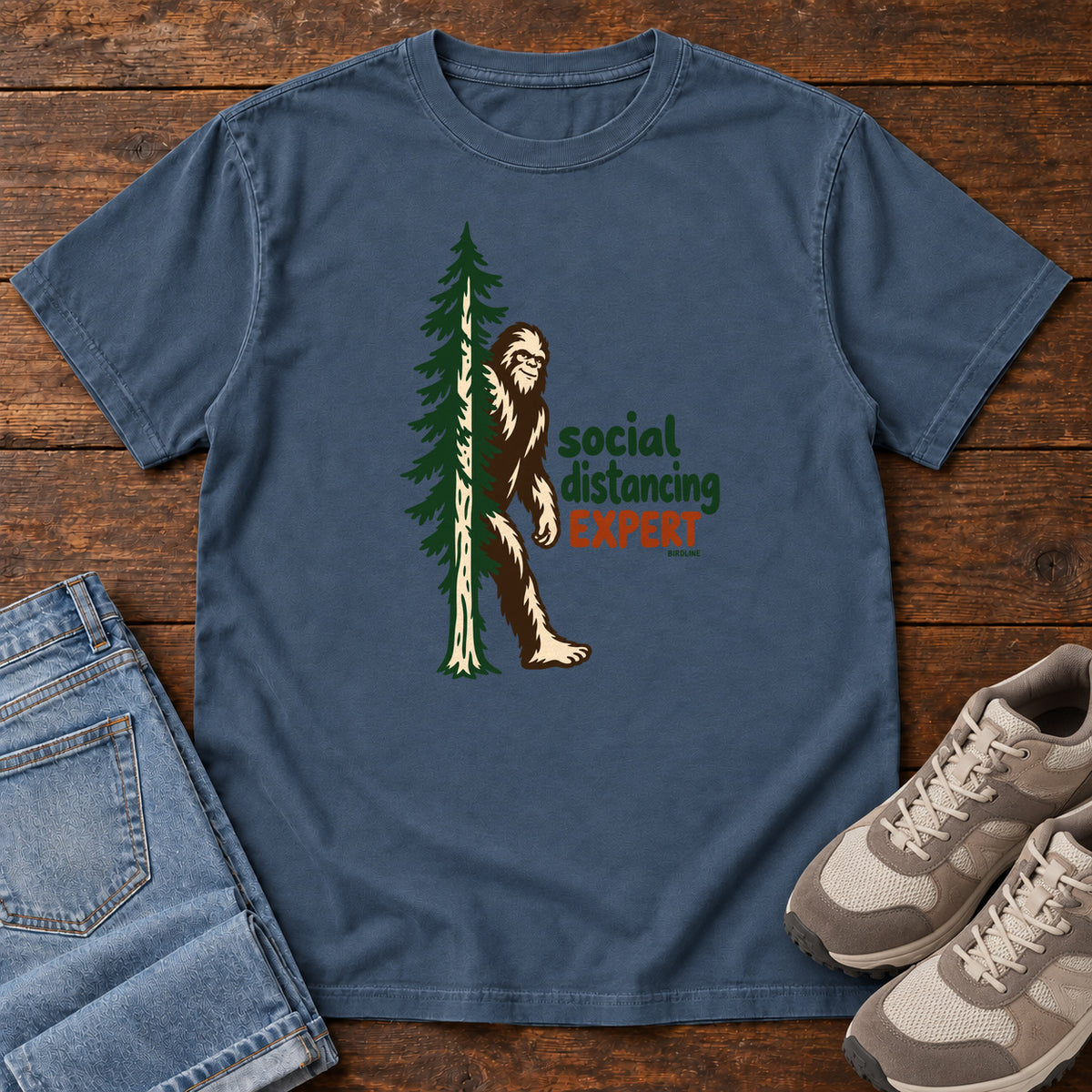Midnight Social Distancing Expert T-Shirt with Bigfoot beside an evergreen and Social Distancing Expert lettering; front flat lay on wood.