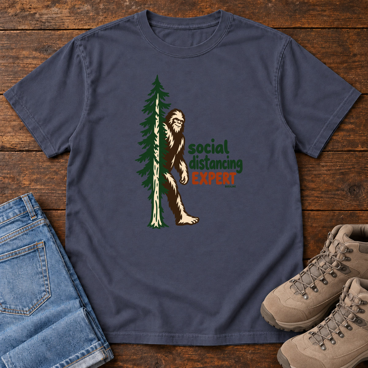 Navy Social Distancing Expert T-Shirt with Bigfoot beside an evergreen and Social Distancing Expert lettering; front flat lay on wood.