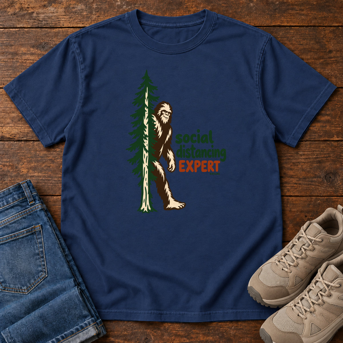 True Navy Social Distancing Expert T-Shirt with Bigfoot beside an evergreen and Social Distancing Expert lettering; front flat lay on wood.