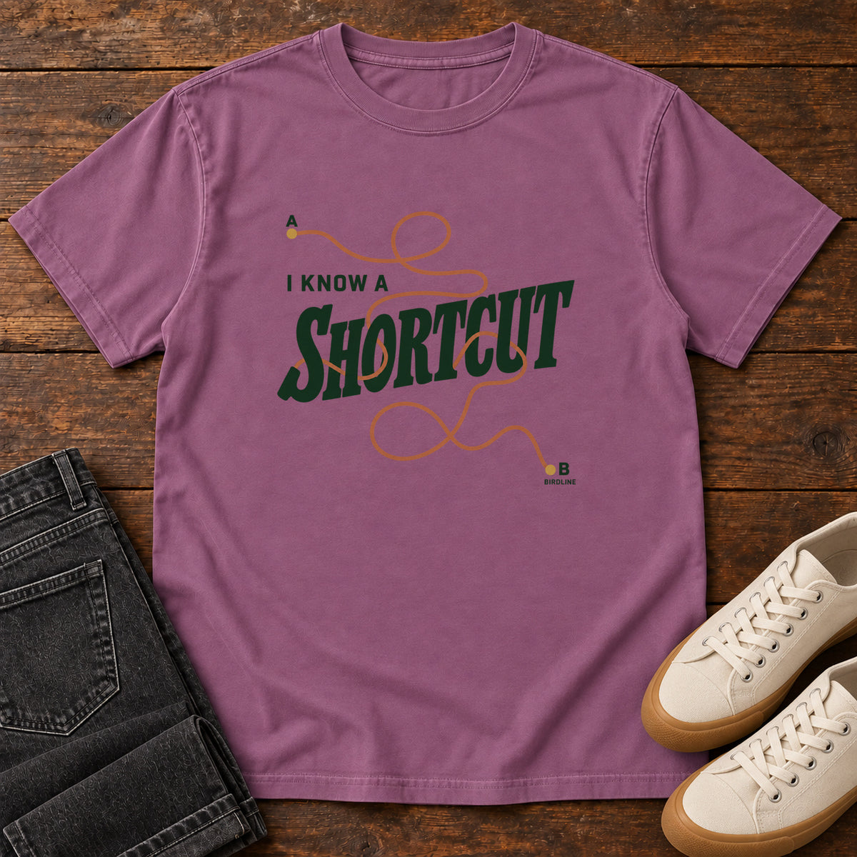 I Know a Shortcut T-Shirt in Berry, front laid flat on wood, featuring dark green slogan lettering crossed by a looping orange route line.