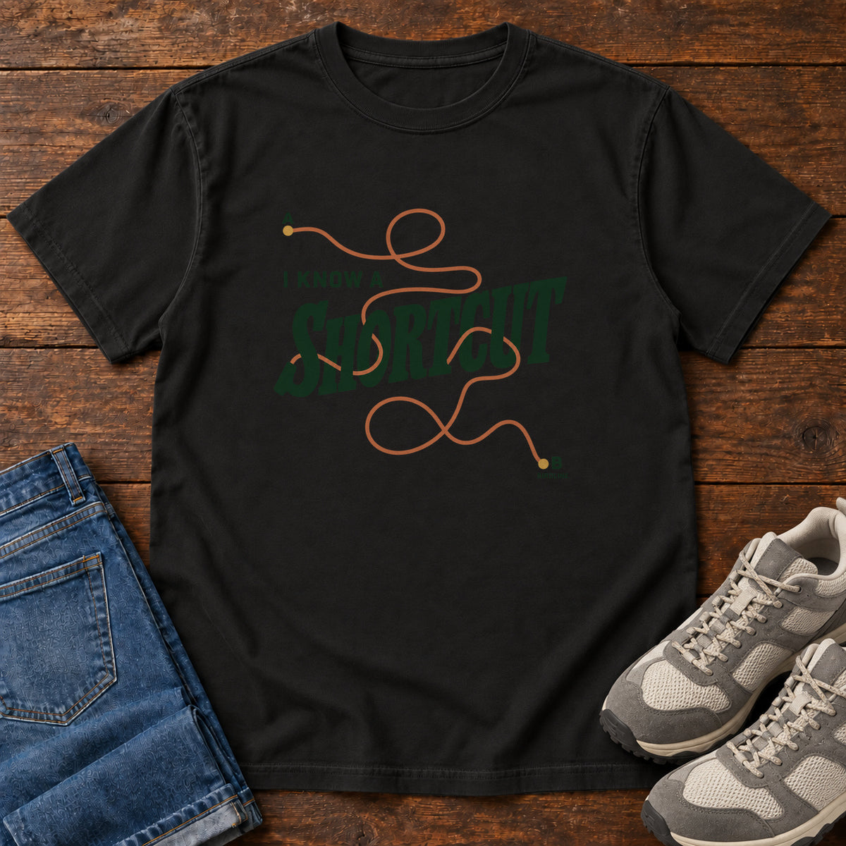 I Know a Shortcut T-Shirt in Black, front laid flat on wood, featuring dark green slogan lettering crossed by a looping orange route line.