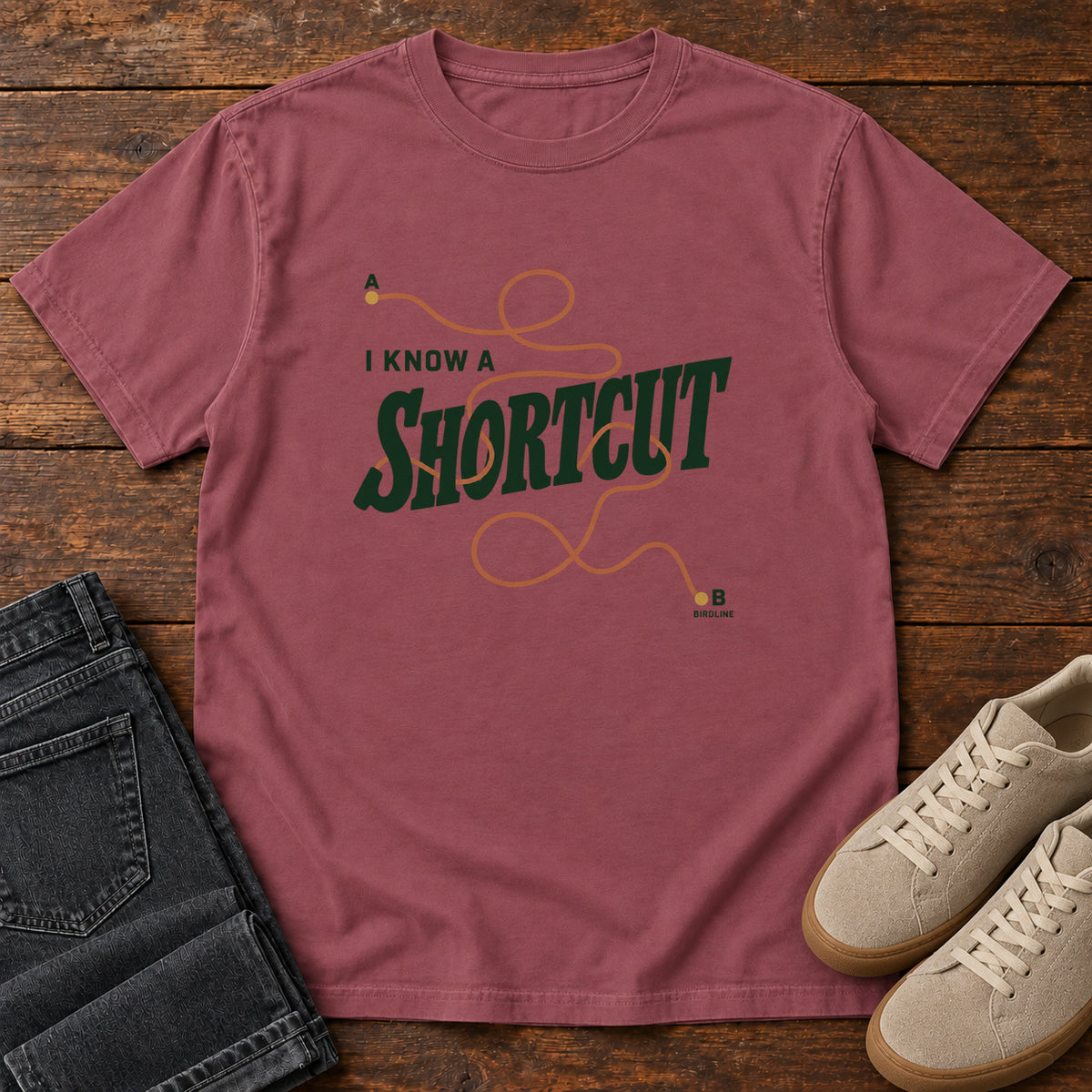 I Know a Shortcut T-Shirt in Brick, front laid flat on wood, featuring dark green slogan lettering crossed by a looping orange route line.
