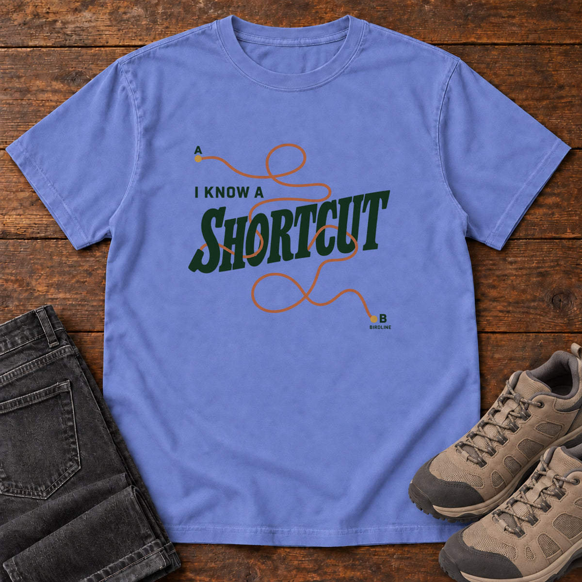 I Know a Shortcut T-Shirt in Flo Blue, front laid flat on wood, featuring dark green slogan lettering crossed by a looping orange route line.