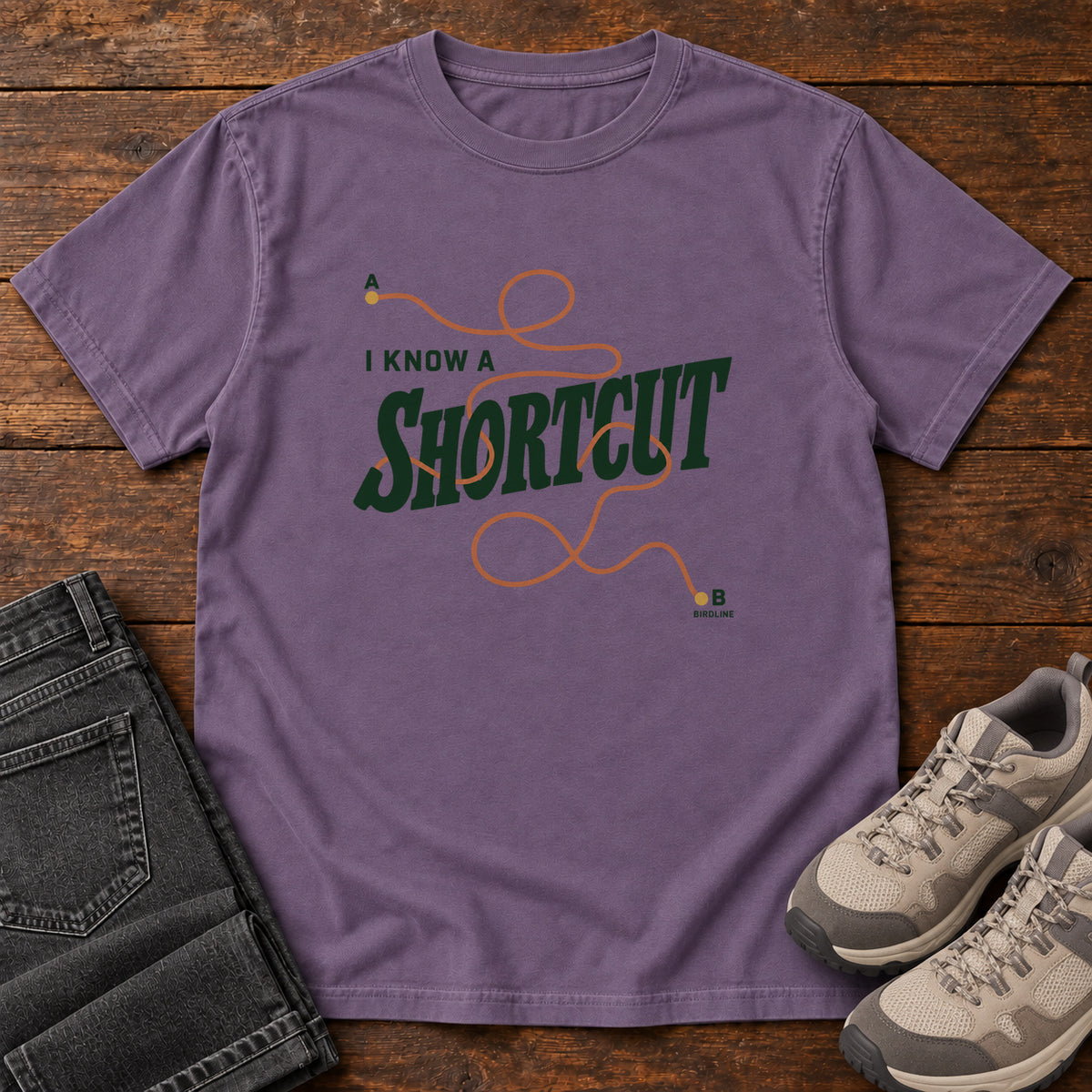 I Know a Shortcut T-Shirt in Grape, front laid flat on wood, featuring dark green slogan lettering crossed by a looping orange route line.