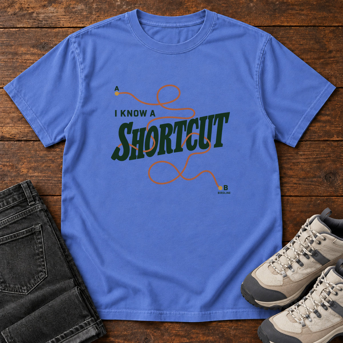 I Know a Shortcut T-Shirt in Mystic Blue, front laid flat on wood, featuring dark green slogan lettering crossed by a looping orange route line.
