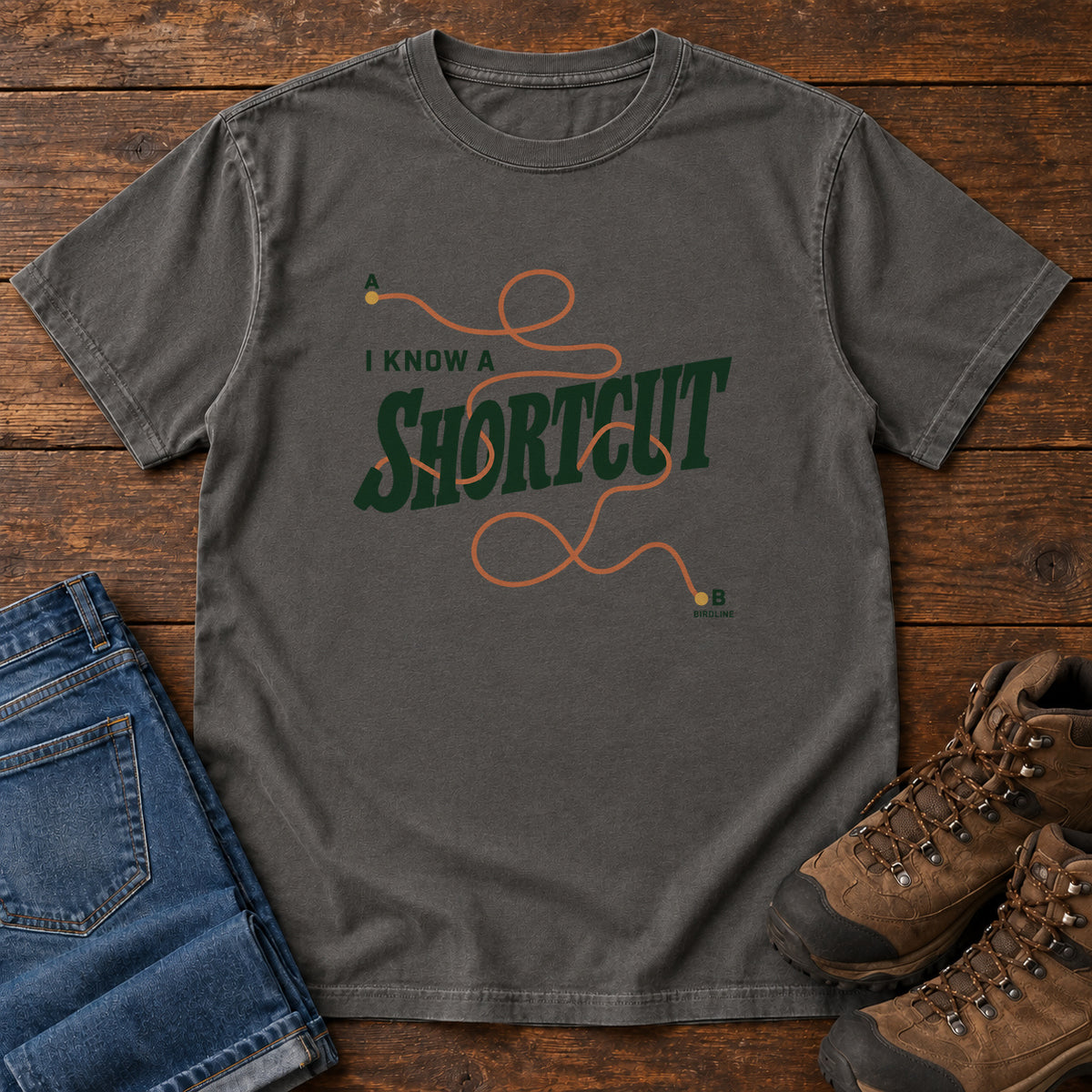 I Know a Shortcut T-Shirt in Pepper, front laid flat on wood, featuring dark green slogan lettering crossed by a looping orange route line.