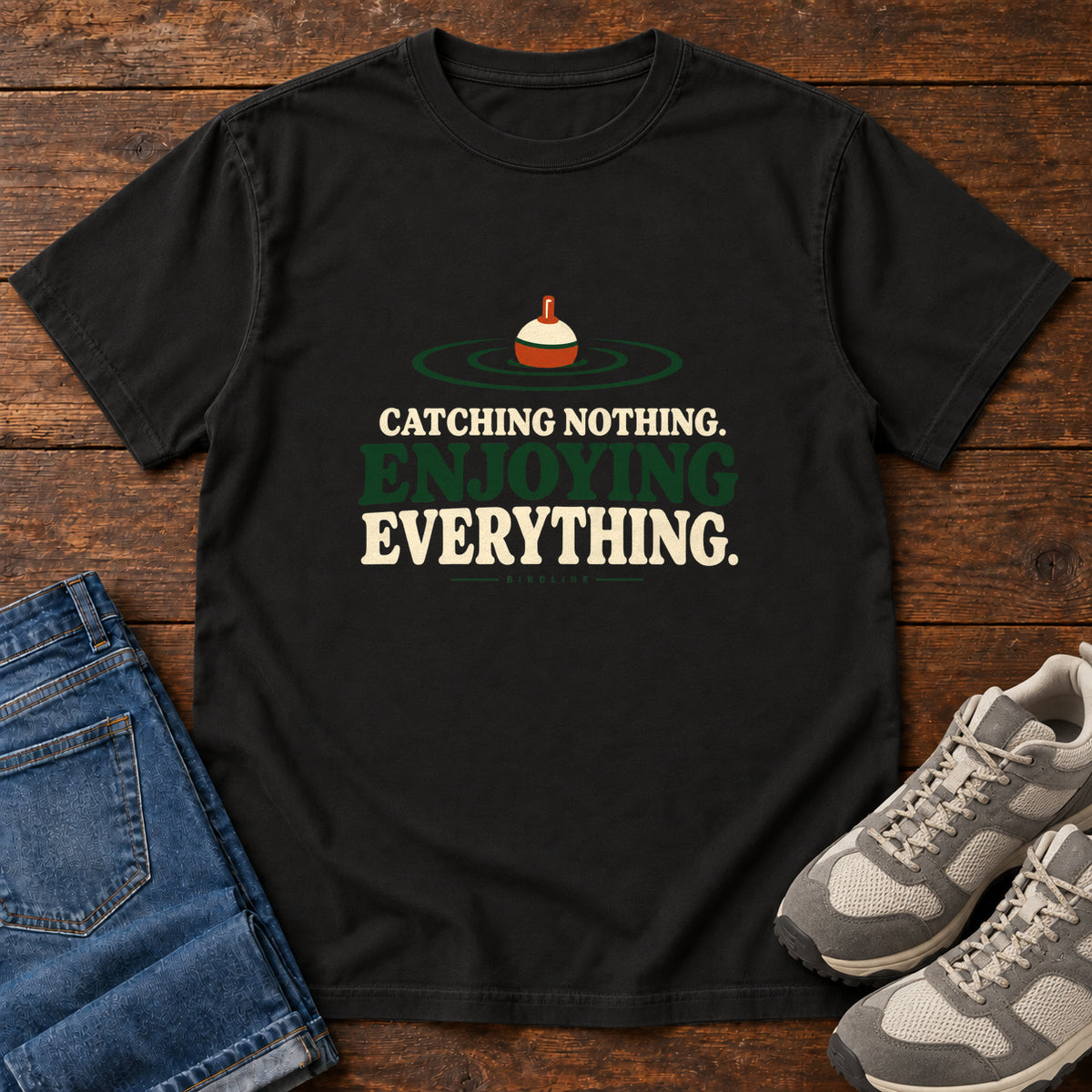 Black Catching Nothing Enjoying Everything T-Shirt with a red-and-cream fishing bobber in green ripples above bold lettering; front flat lay on wood.