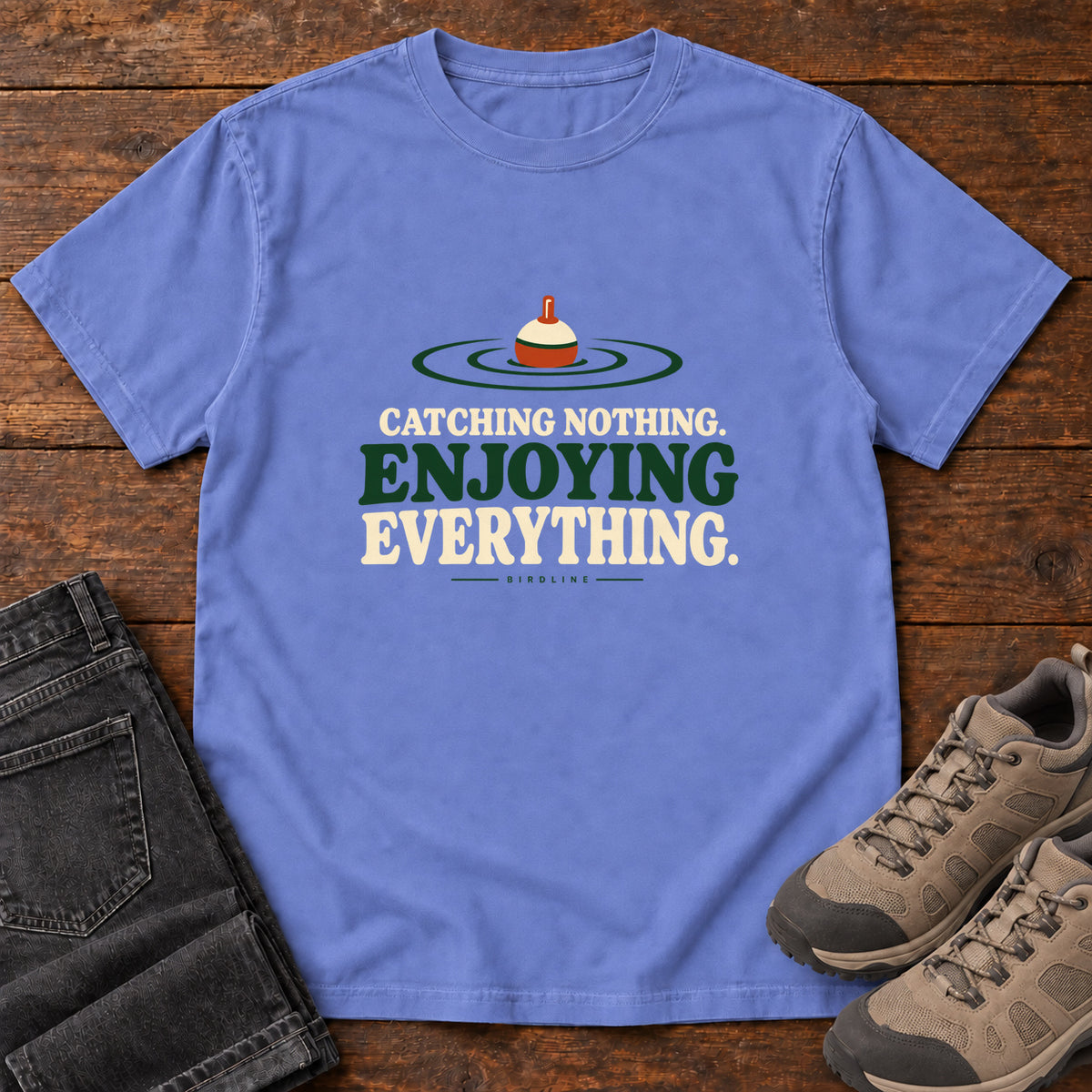 Flo Blue Catching Nothing Enjoying Everything T-Shirt with a red-and-cream fishing bobber in green ripples above bold lettering; front flat lay on wood.