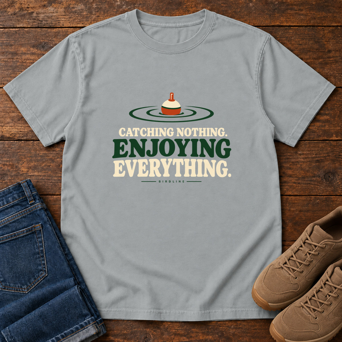 Granite Catching Nothing Enjoying Everything T-Shirt with a red-and-cream fishing bobber in green ripples above bold lettering; front flat lay on wood.