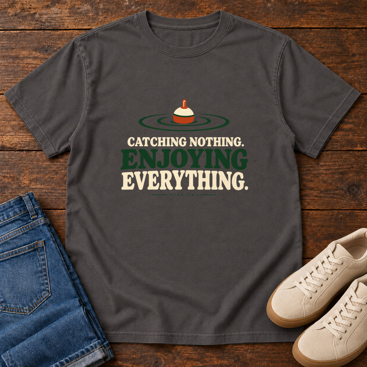 Graphite Catching Nothing Enjoying Everything T-Shirt with a red-and-cream fishing bobber in green ripples above bold lettering; front flat lay on wood.