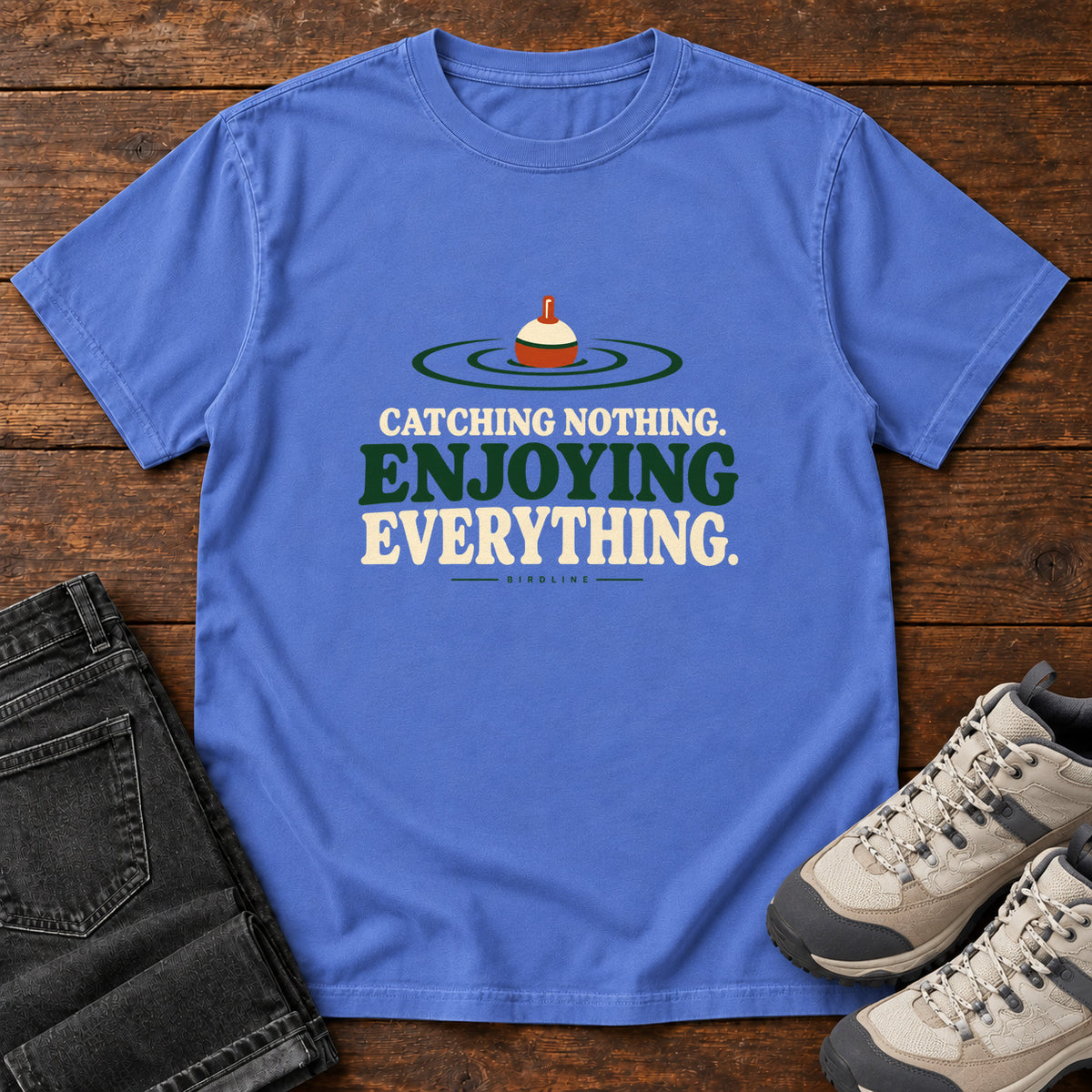 Mystic Blue Catching Nothing Enjoying Everything T-Shirt with a red-and-cream fishing bobber in green ripples above bold lettering; front flat lay on wood.