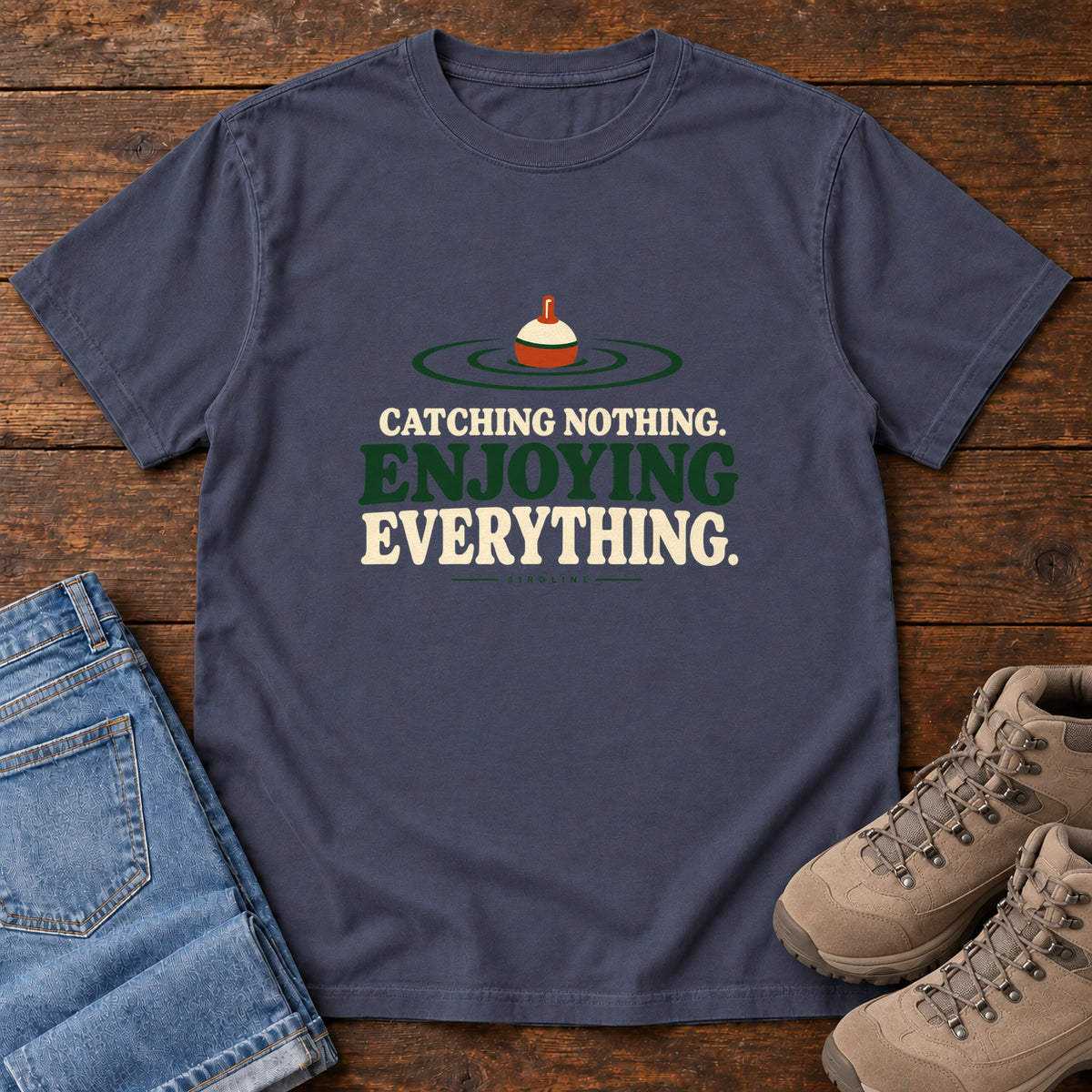 Navy Catching Nothing Enjoying Everything T-Shirt with a red-and-cream fishing bobber in green ripples above bold lettering; front flat lay on wood.
