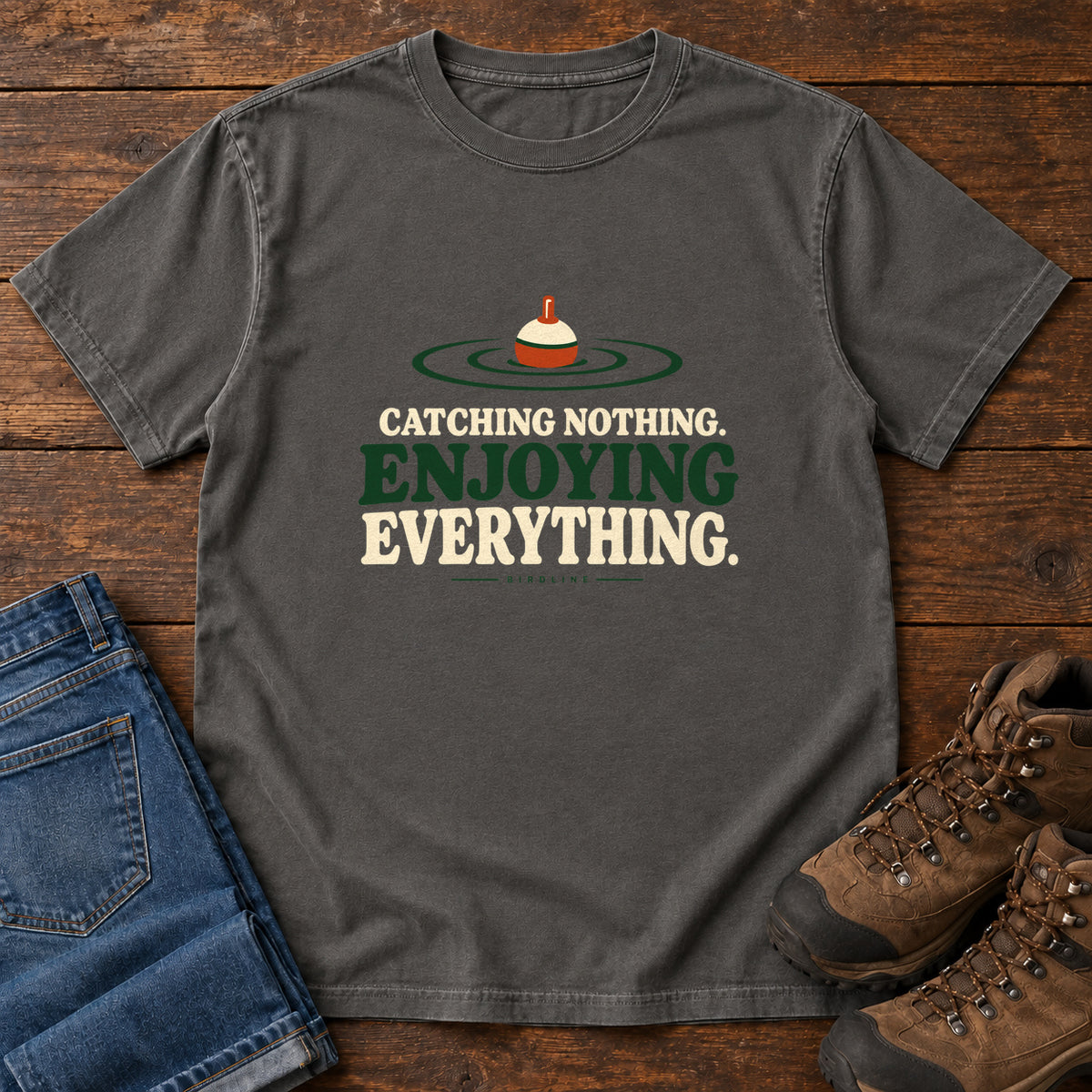 Pepper Catching Nothing Enjoying Everything T-Shirt with a red-and-cream fishing bobber in green ripples above bold lettering; front flat lay on wood.