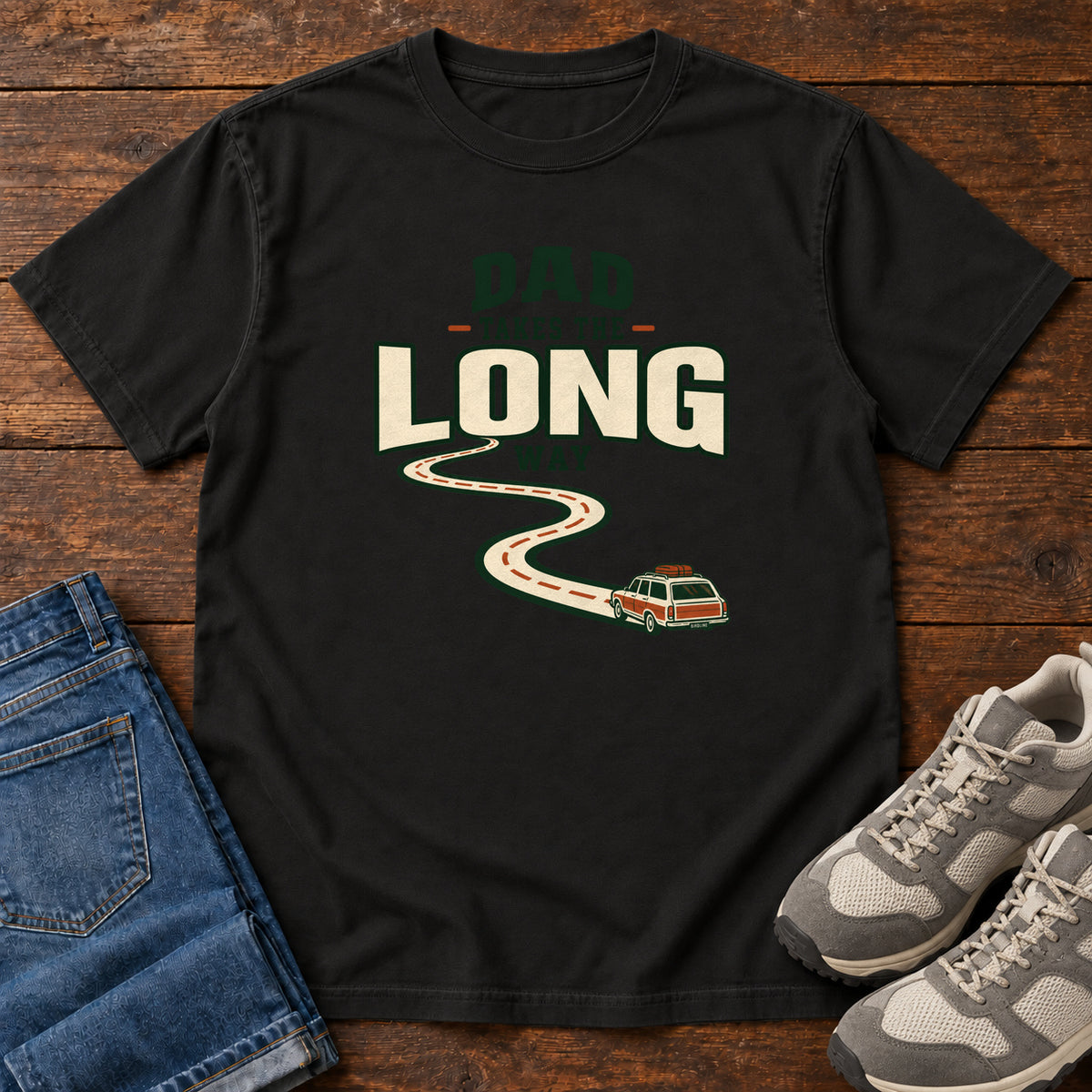Black Dad Takes the Long Way T-Shirt with a winding road leading to a station wagon beneath bold cream and green lettering, front flat lay on wood.