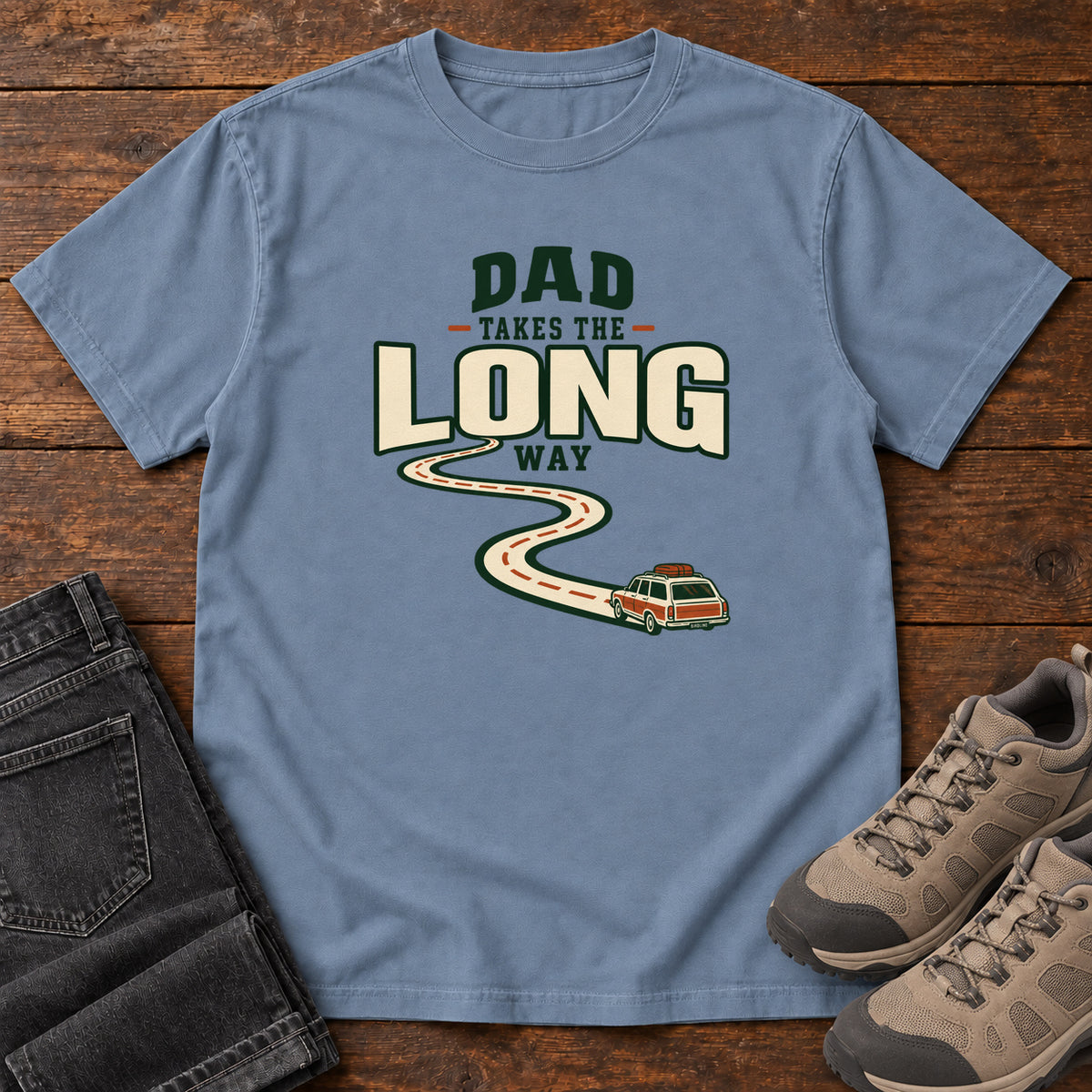 Blue Jean Dad Takes the Long Way T-Shirt with a winding road leading to a station wagon beneath bold cream and green lettering, front flat lay on wood.