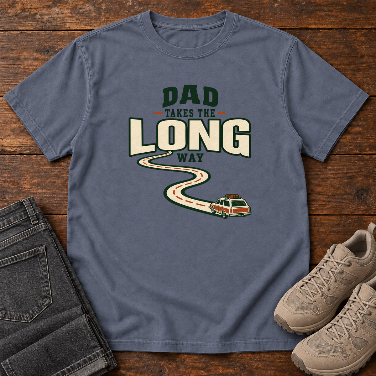 Denim Dad Takes the Long Way T-Shirt with a winding road leading to a station wagon beneath bold cream and green lettering, front flat lay on wood.