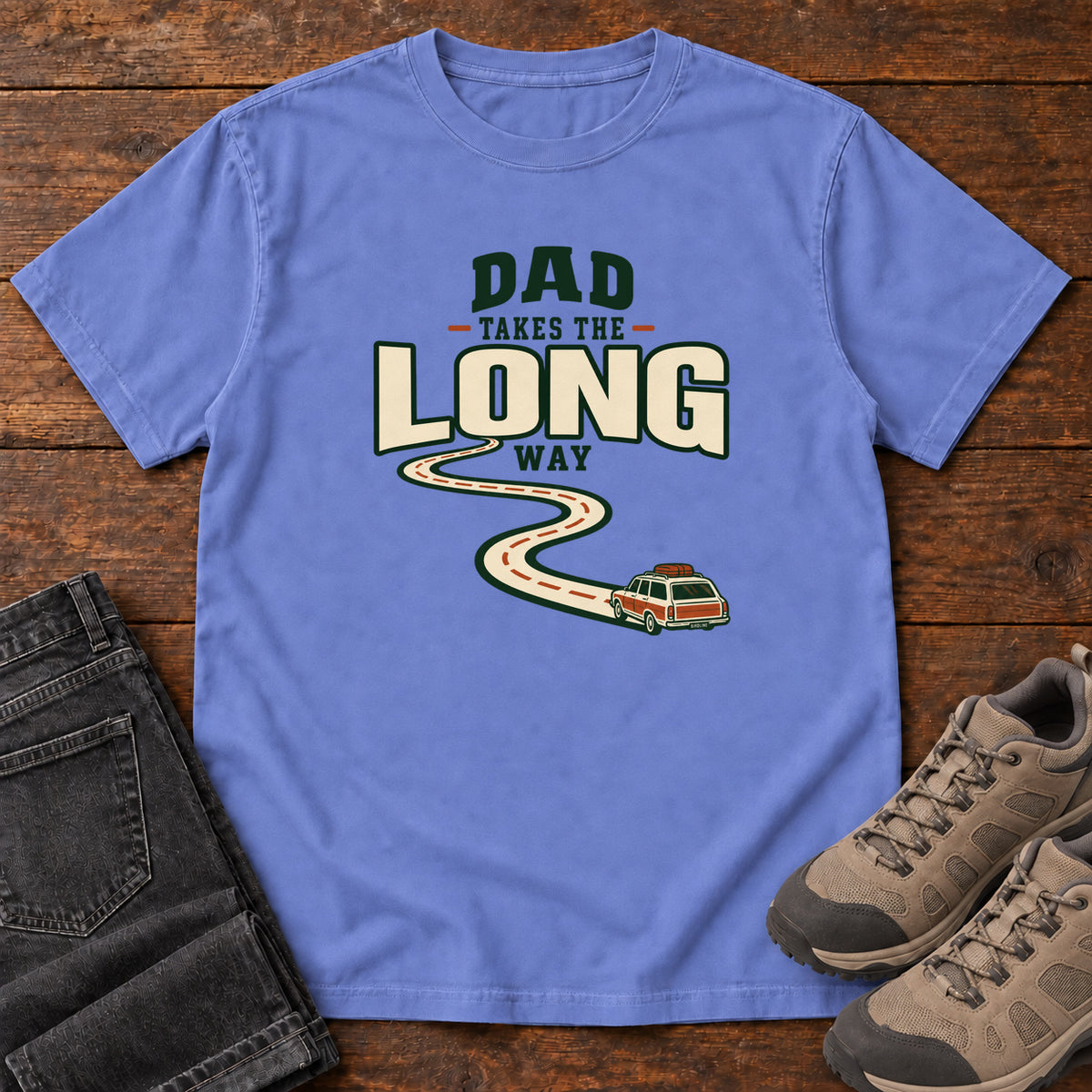 Flo Blue Dad Takes the Long Way T-Shirt with a winding road leading to a station wagon beneath bold cream and green lettering, front flat lay on wood.