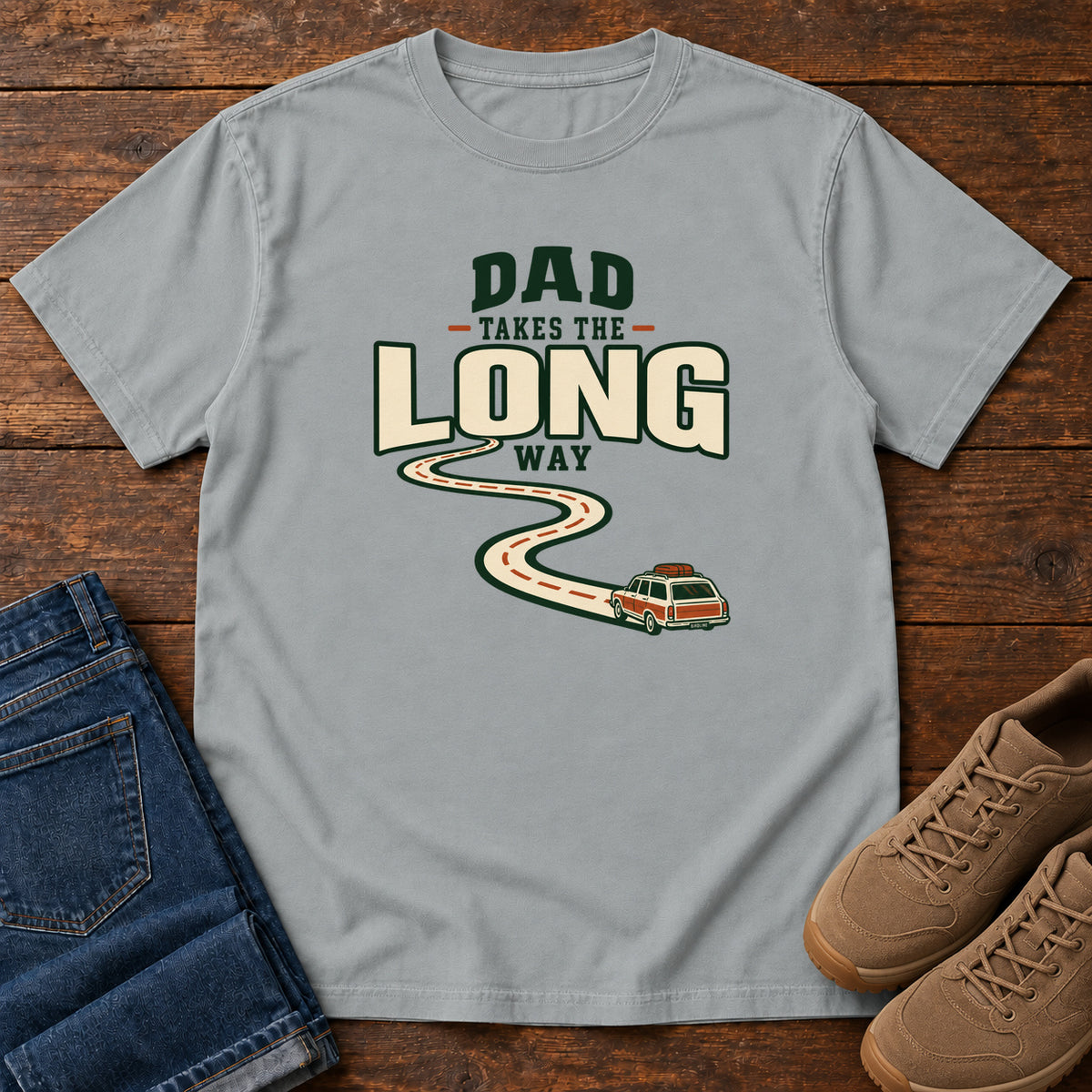 Granite Dad Takes the Long Way T-Shirt with a winding road leading to a station wagon beneath bold cream and green lettering, front flat lay on wood.