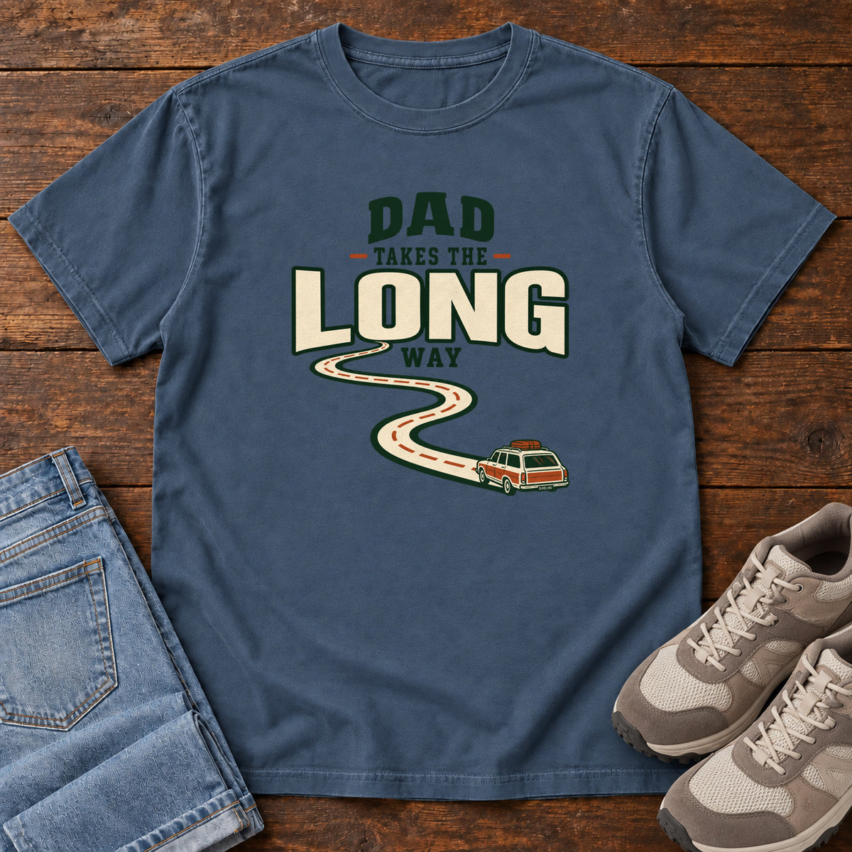 Midnight Dad Takes the Long Way T-Shirt with a winding road leading to a station wagon beneath bold cream and green lettering, front flat lay on wood.