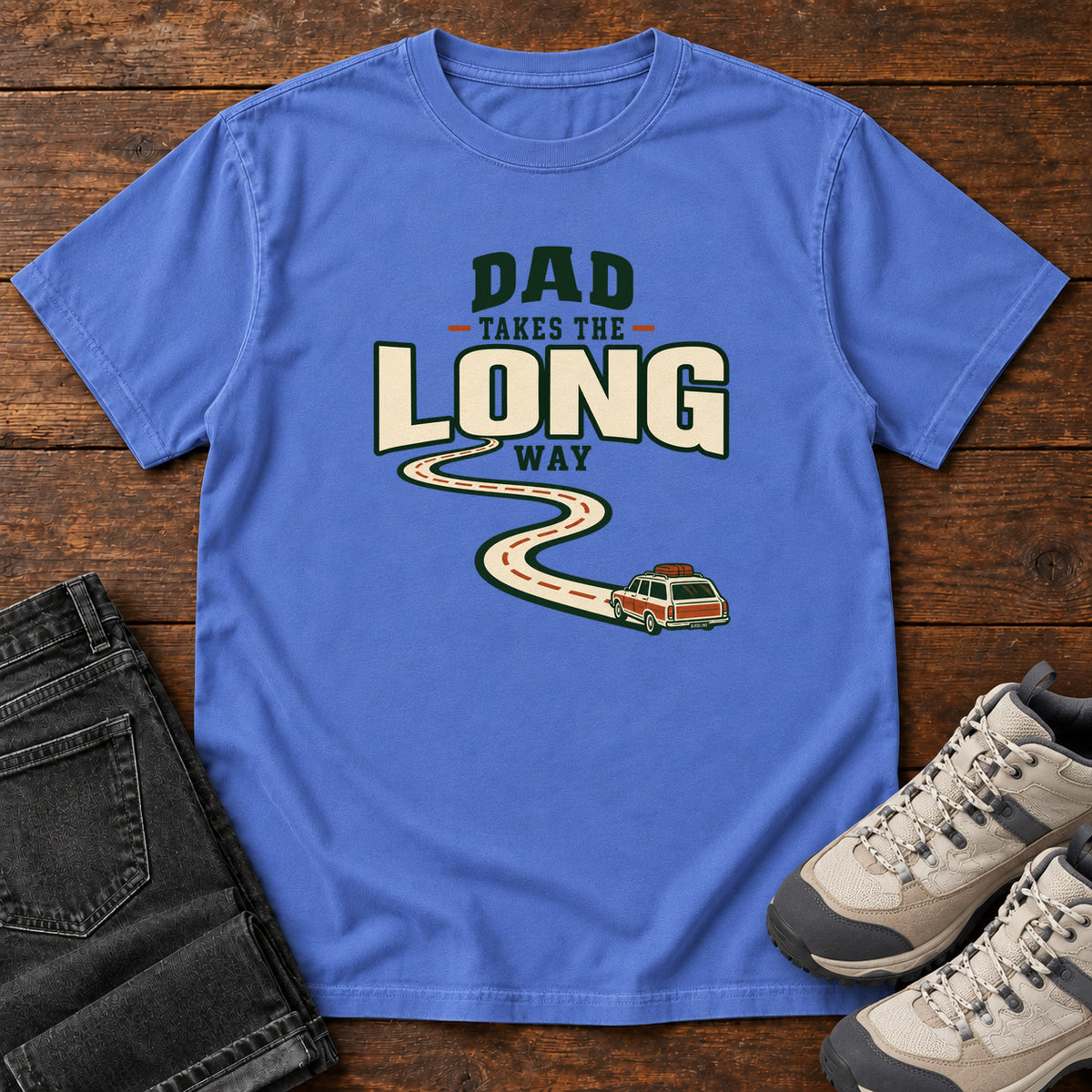 Mystic Blue Dad Takes the Long Way T-Shirt with a winding road leading to a station wagon beneath bold cream and green lettering, front flat lay on wood.