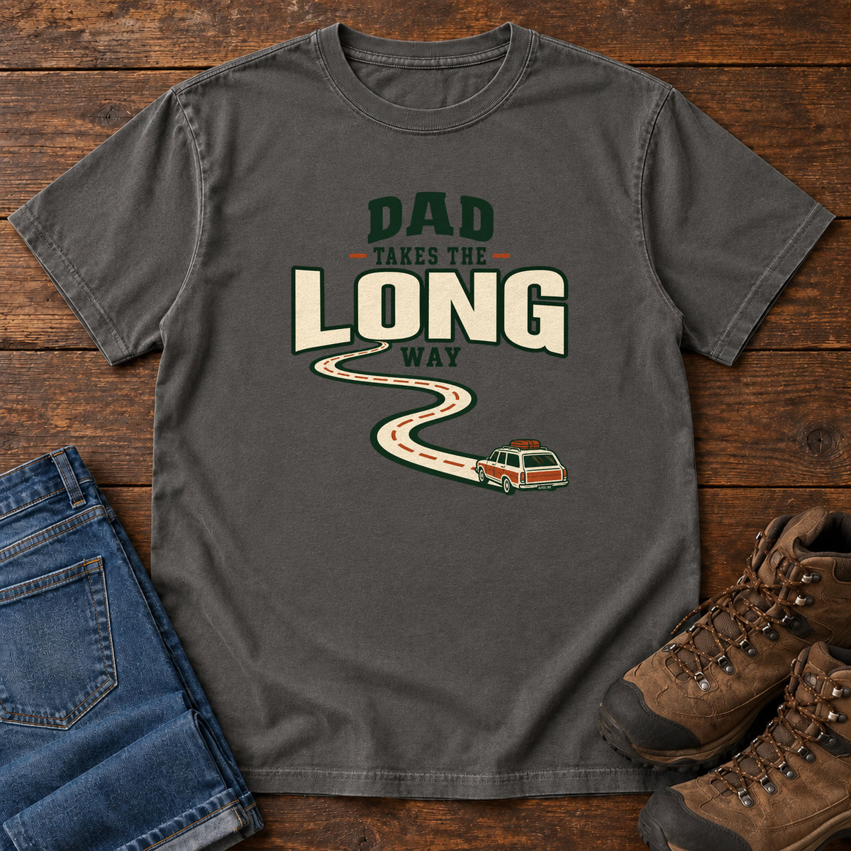 Pepper Dad Takes the Long Way T-Shirt with a winding road leading to a station wagon beneath bold cream and green lettering, front flat lay on wood.