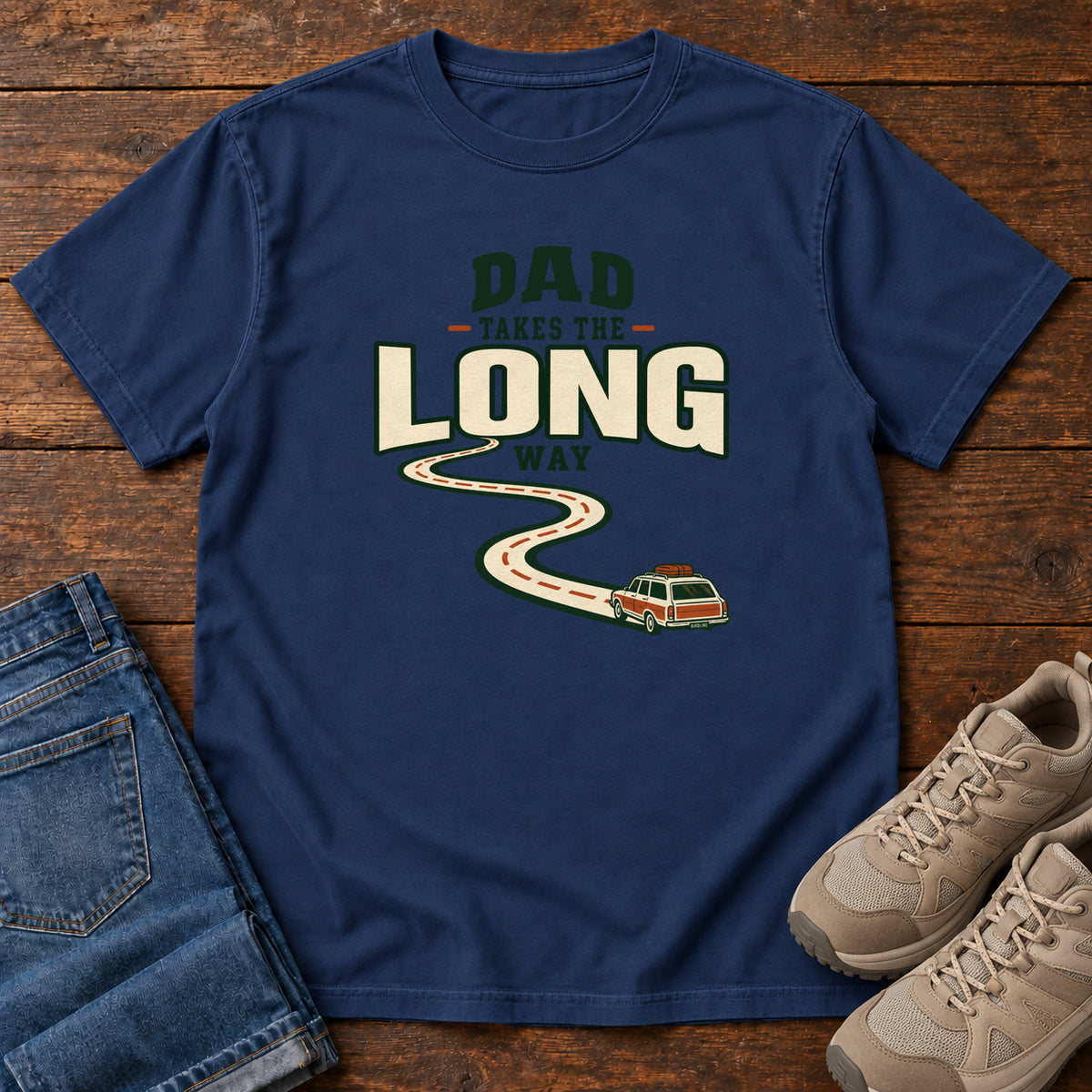 True Navy Dad Takes the Long Way T-Shirt with a winding road leading to a station wagon beneath bold cream and green lettering, front flat lay on wood.