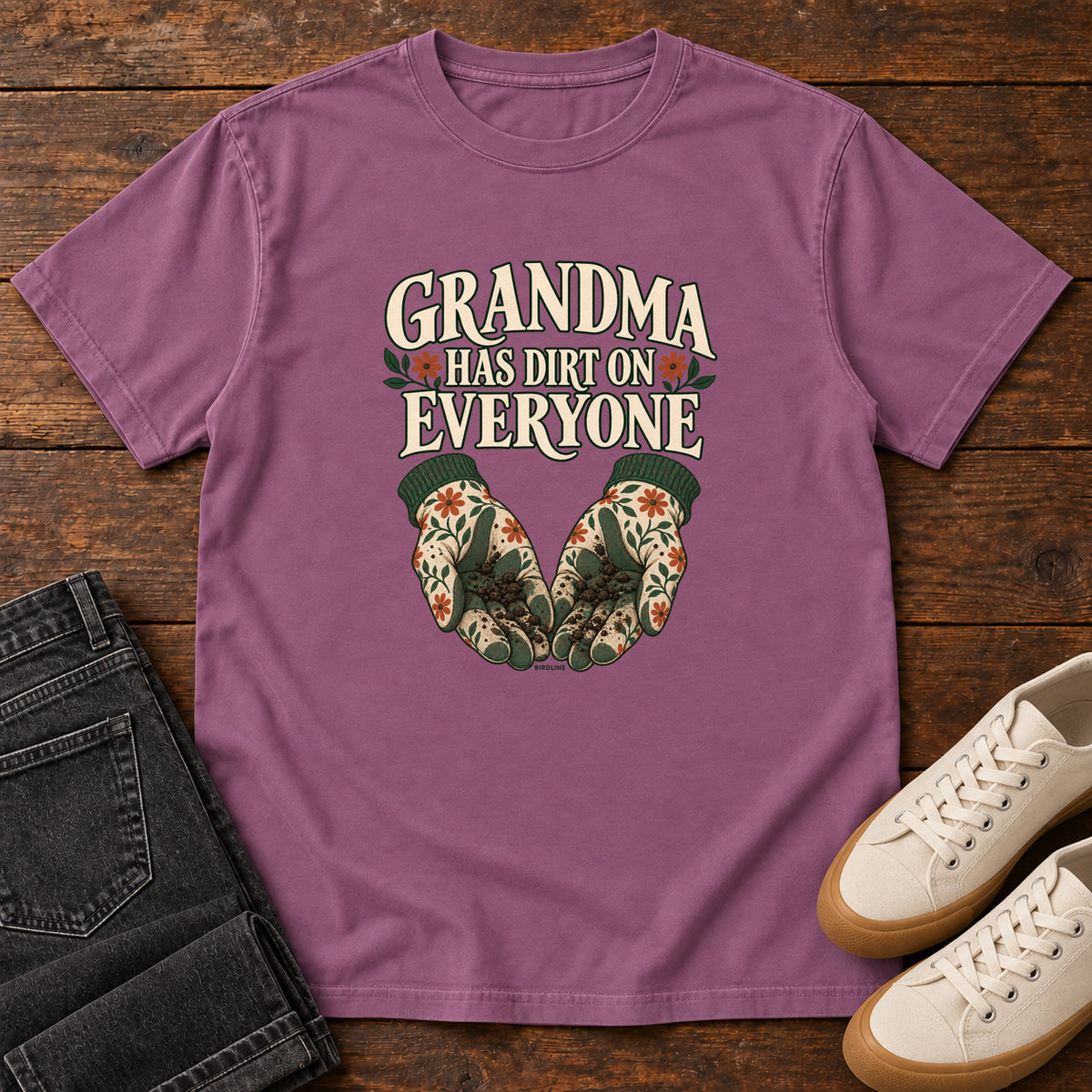 Grandma Has Dirt on Everyone T-Shirt in Berry, front laid flat on wood, featuring floral gardening gloves cupping soil beneath large cream slogan lettering.