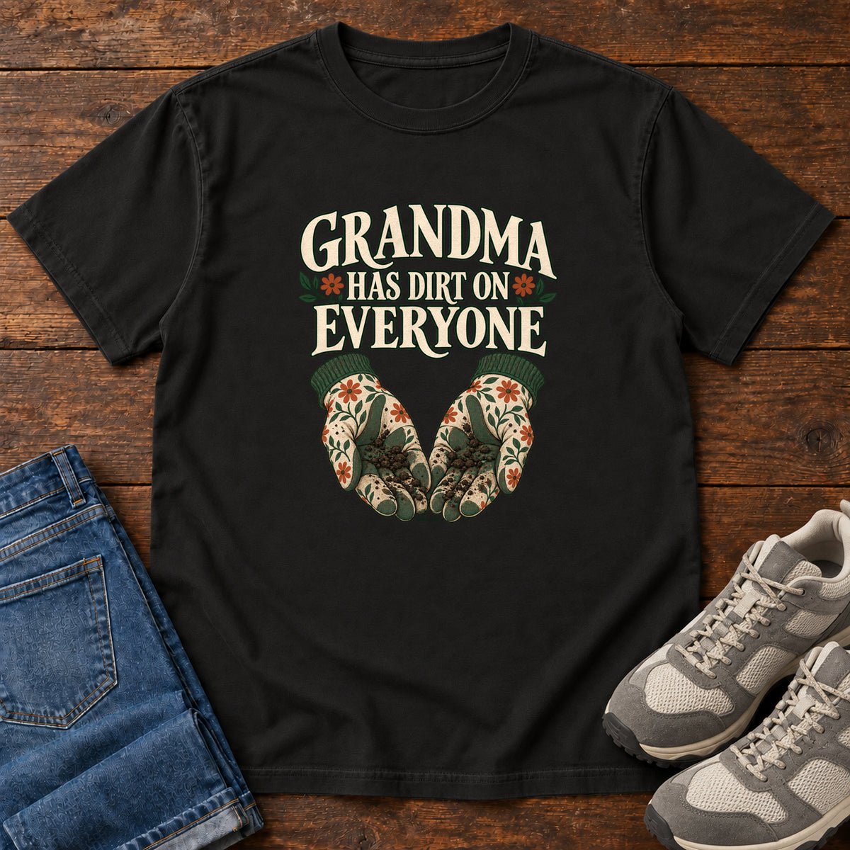 Grandma Has Dirt on Everyone T-Shirt in Black, front laid flat on wood, featuring floral gardening gloves cupping soil beneath large cream slogan lettering.