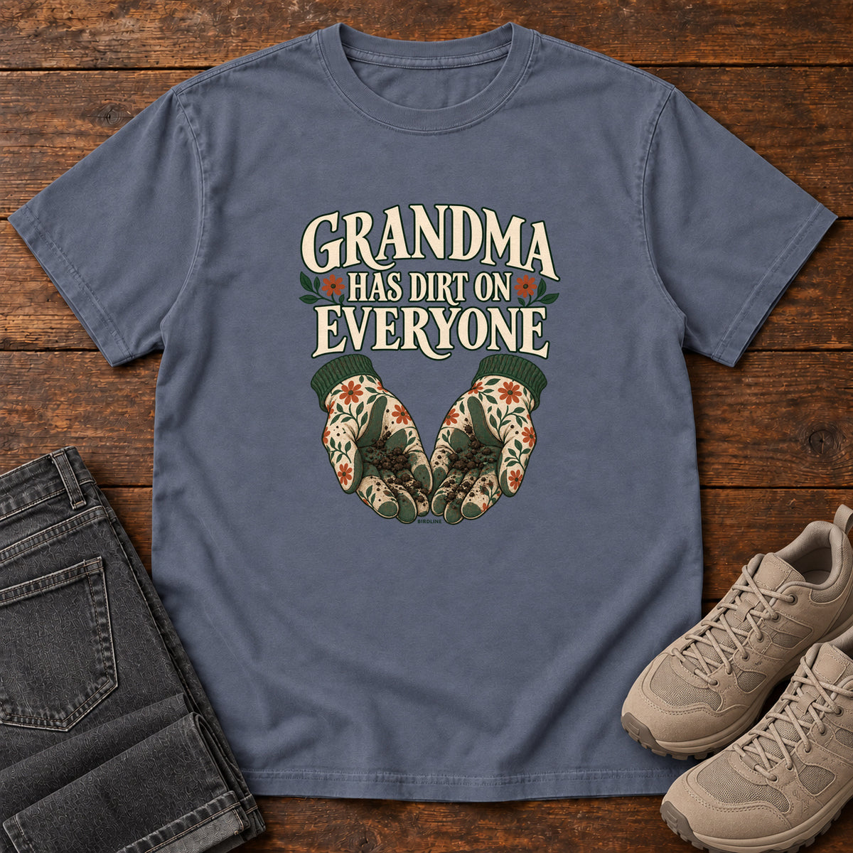 Grandma Has Dirt on Everyone T-Shirt in Denim, front laid flat on wood, featuring floral gardening gloves cupping soil beneath large cream slogan lettering.