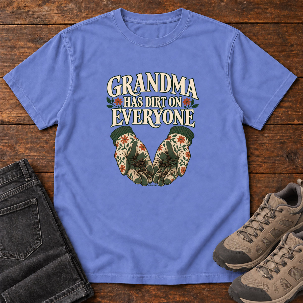 Grandma Has Dirt on Everyone T-Shirt in Flo Blue, front laid flat on wood, featuring floral gardening gloves cupping soil beneath large cream slogan lettering.