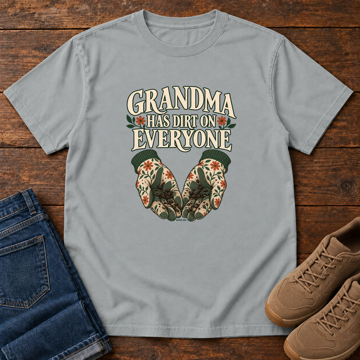 Grandma Has Dirt on Everyone T-Shirt in Granite, front laid flat on wood, featuring floral gardening gloves cupping soil beneath large cream slogan lettering.