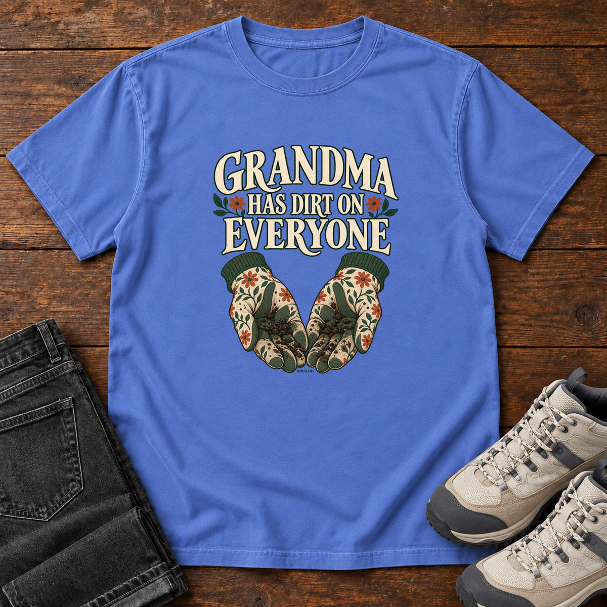 Grandma Has Dirt on Everyone T-Shirt in Mystic Blue, front laid flat on wood, featuring floral gardening gloves cupping soil beneath large cream slogan lettering.