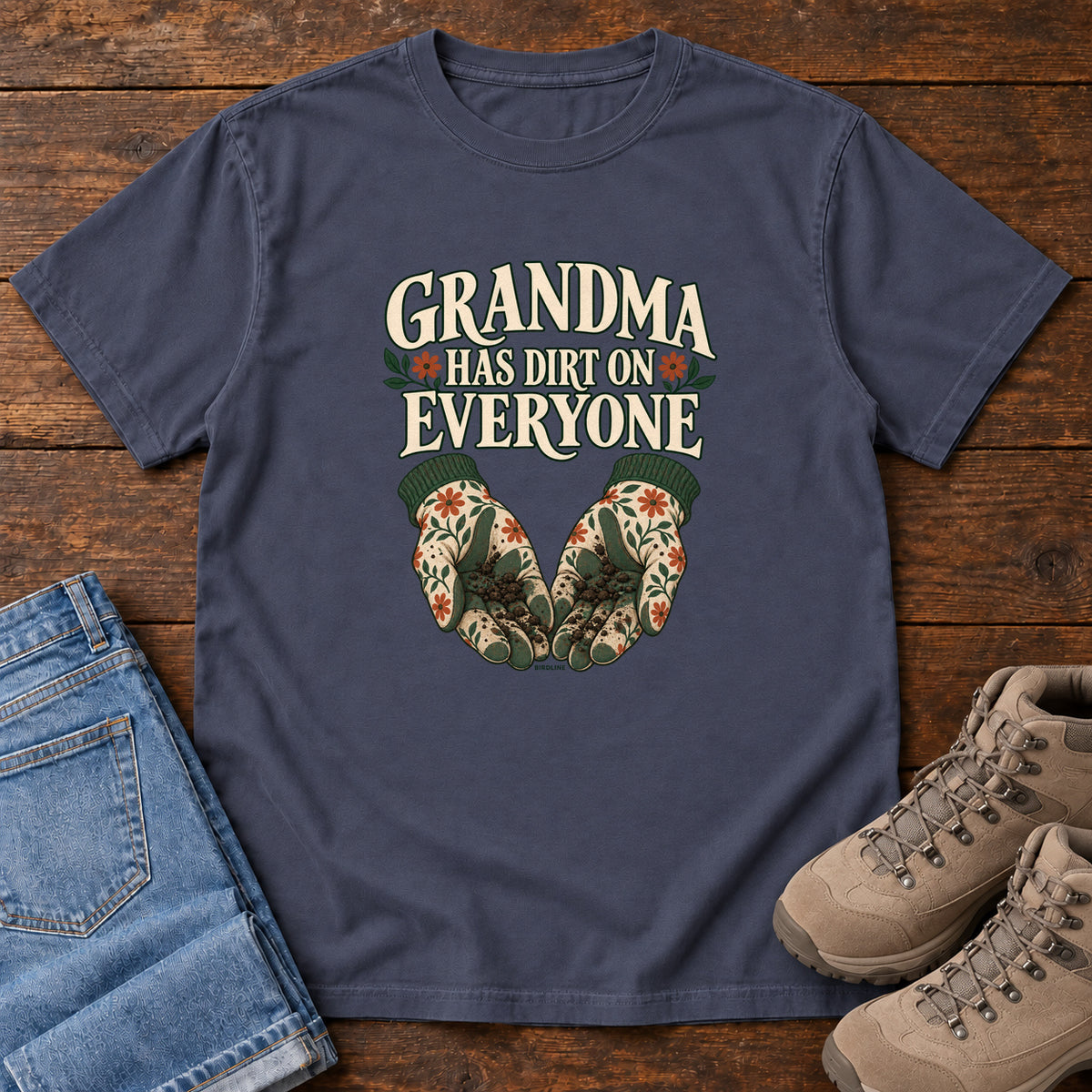 Grandma Has Dirt on Everyone T-Shirt in Navy, front laid flat on wood, featuring floral gardening gloves cupping soil beneath large cream slogan lettering.