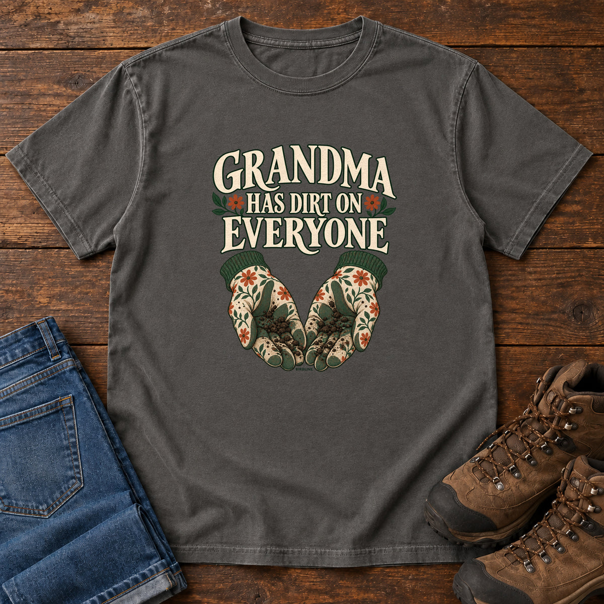 Grandma Has Dirt on Everyone T-Shirt in Pepper, front laid flat on wood, featuring floral gardening gloves cupping soil beneath large cream slogan lettering.