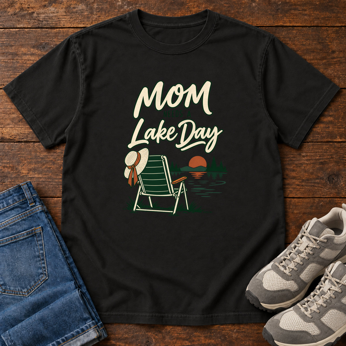 Black Mom Needs a Lake Day T-Shirt with a striped folding chair, sunhat and lakeside sunset illustration, front flat lay on wood.